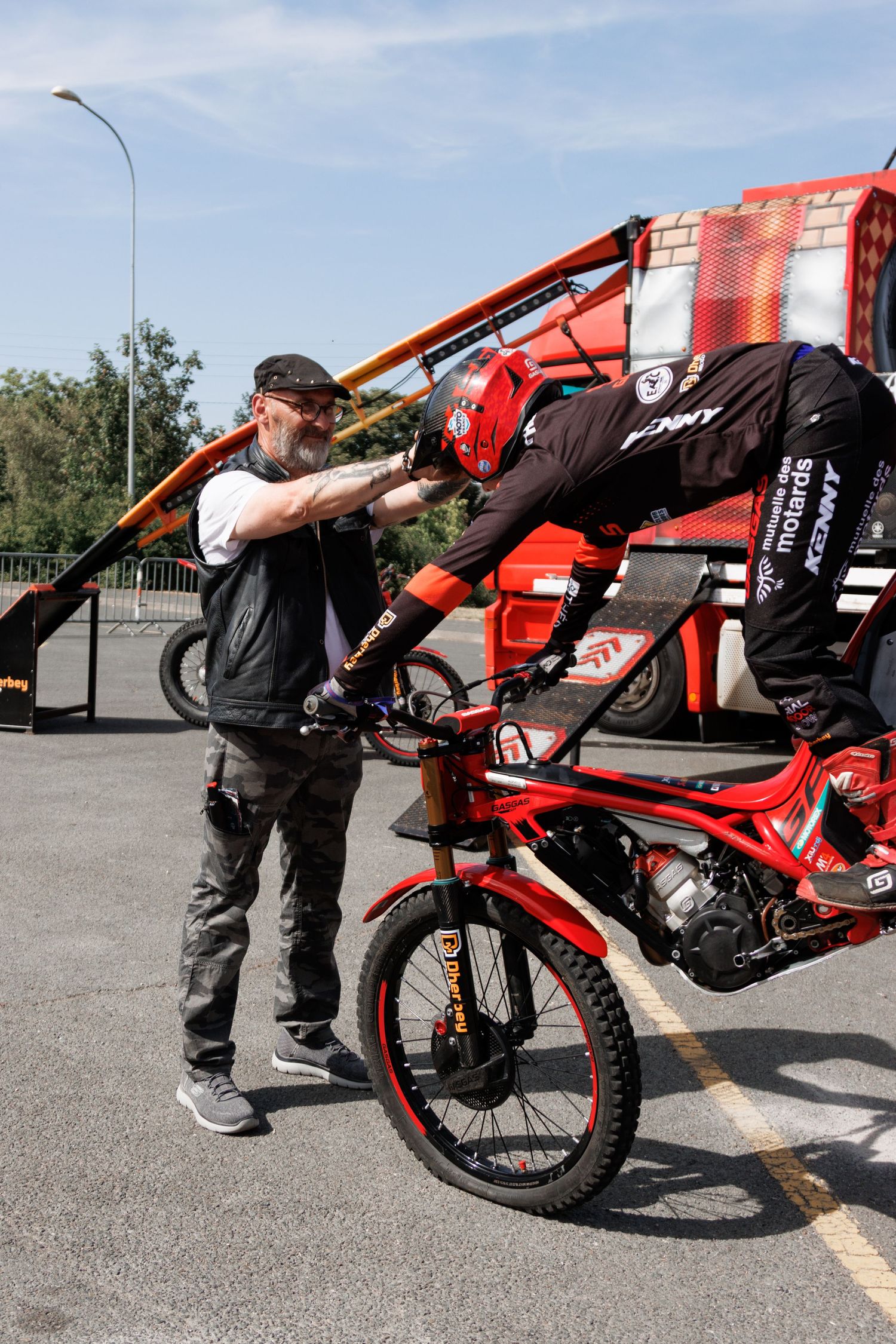 A rider in a red helmet leans over a red motorcycle while a bearded man assists during an outdoor motorsport event.