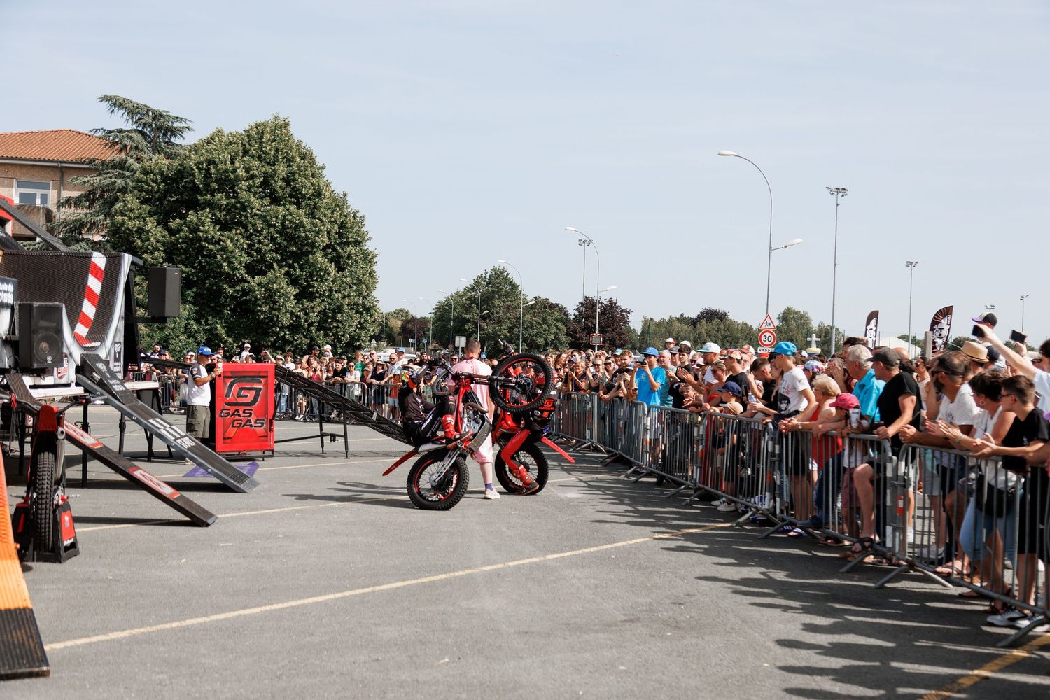 Motorcycle stunt rider performs wheelie for large crowd at outdoor motorsport event on sunny day.