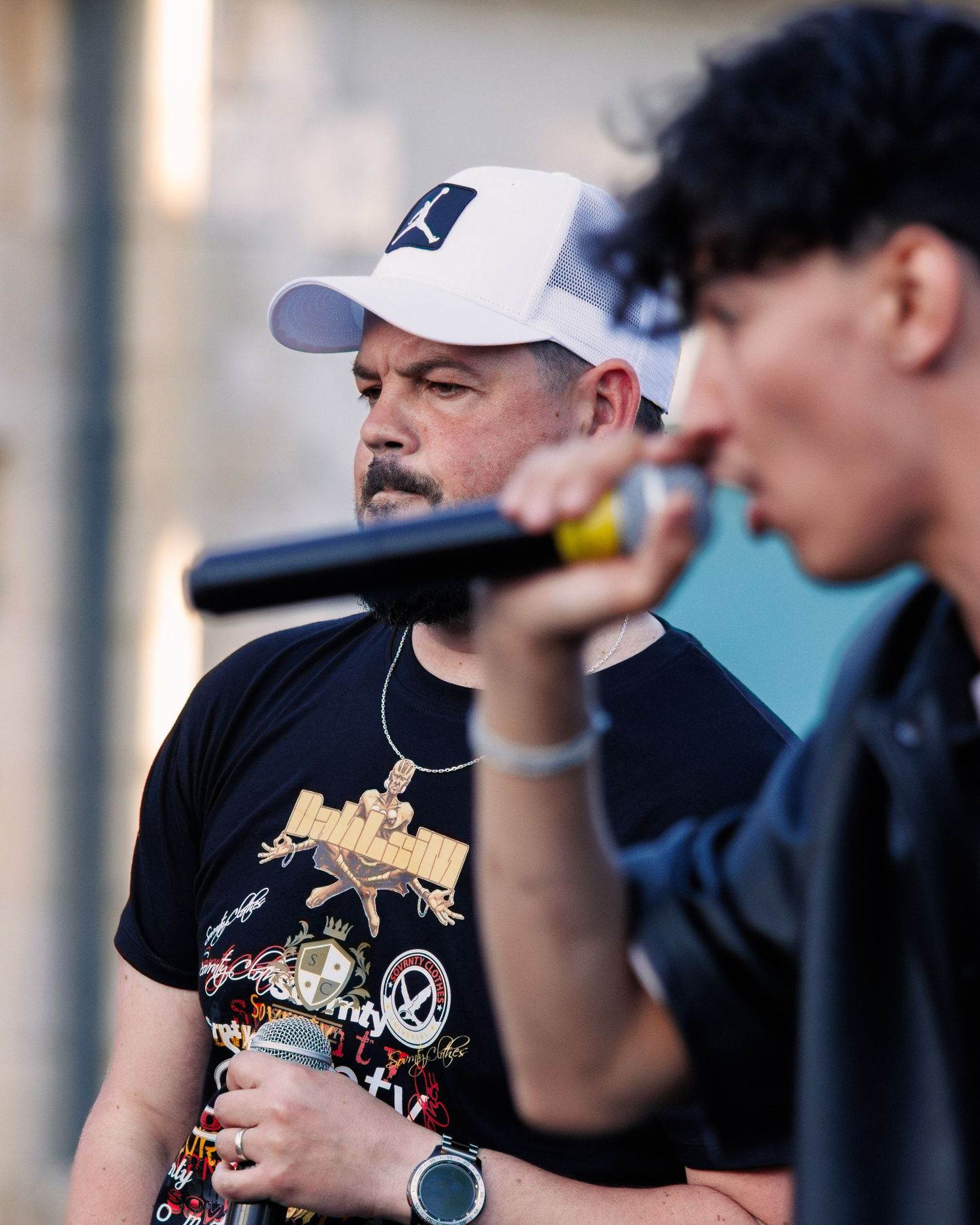 Two performers on stage singing into microphones, wearing baseball caps and casual attire.