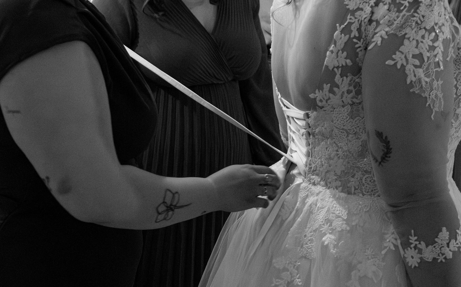 Black and white photo of a bridesmaid lacing up the back of a bride's lace wedding dress.