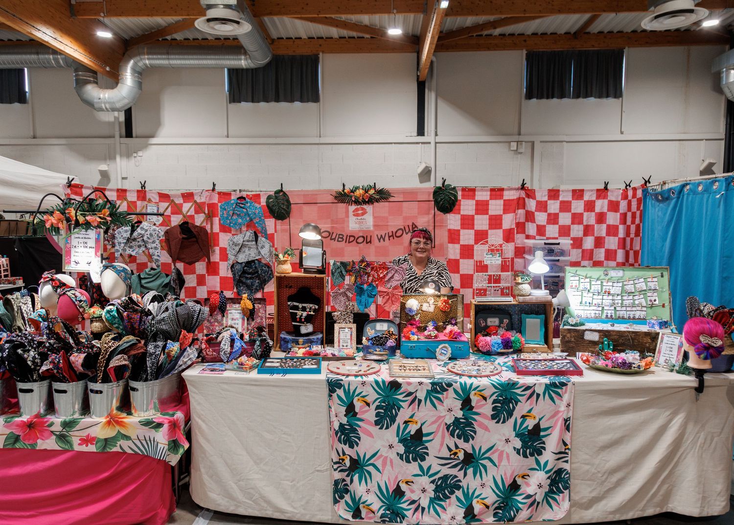Colorful craft fair booth displaying tropical-themed items, pink and red checkered backdrop, vibrant tablecloths, and handmade goods.