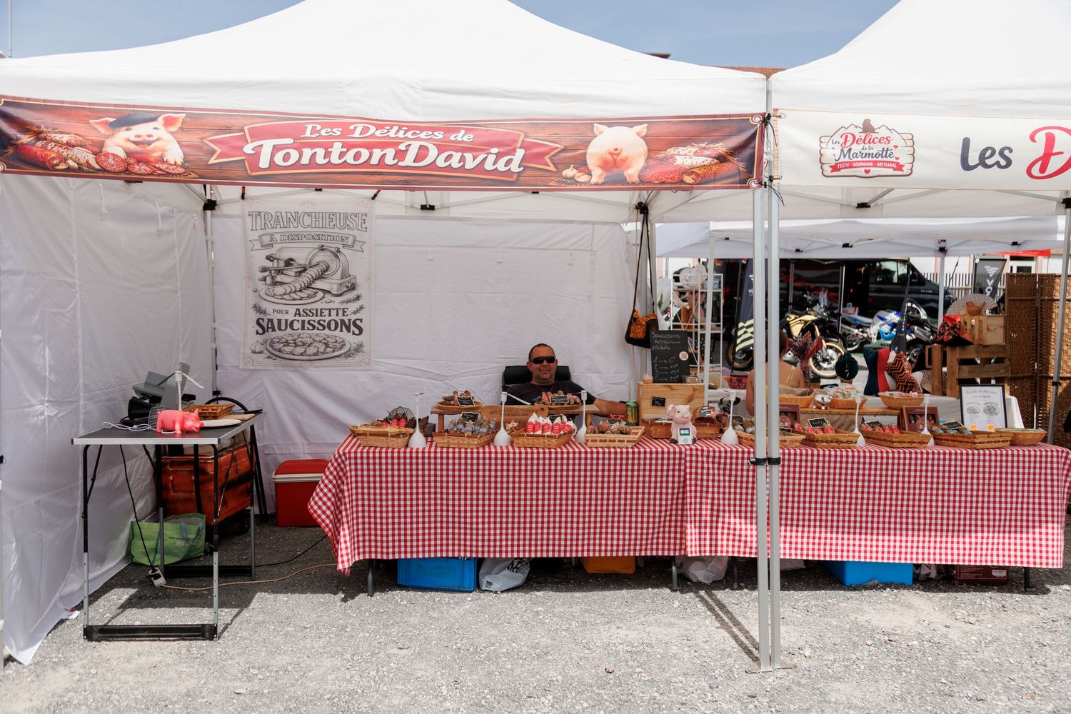 Tonton David French market stall with red checkered tablecloths displaying artisan food products under a branded tent.