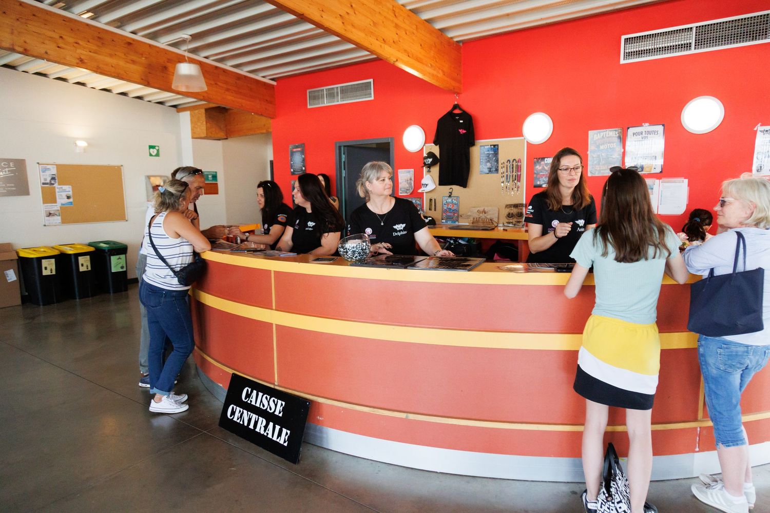 Visitors at a curved red and yellow reception desk labeled Caisse Centrale in a bright French visitor center.