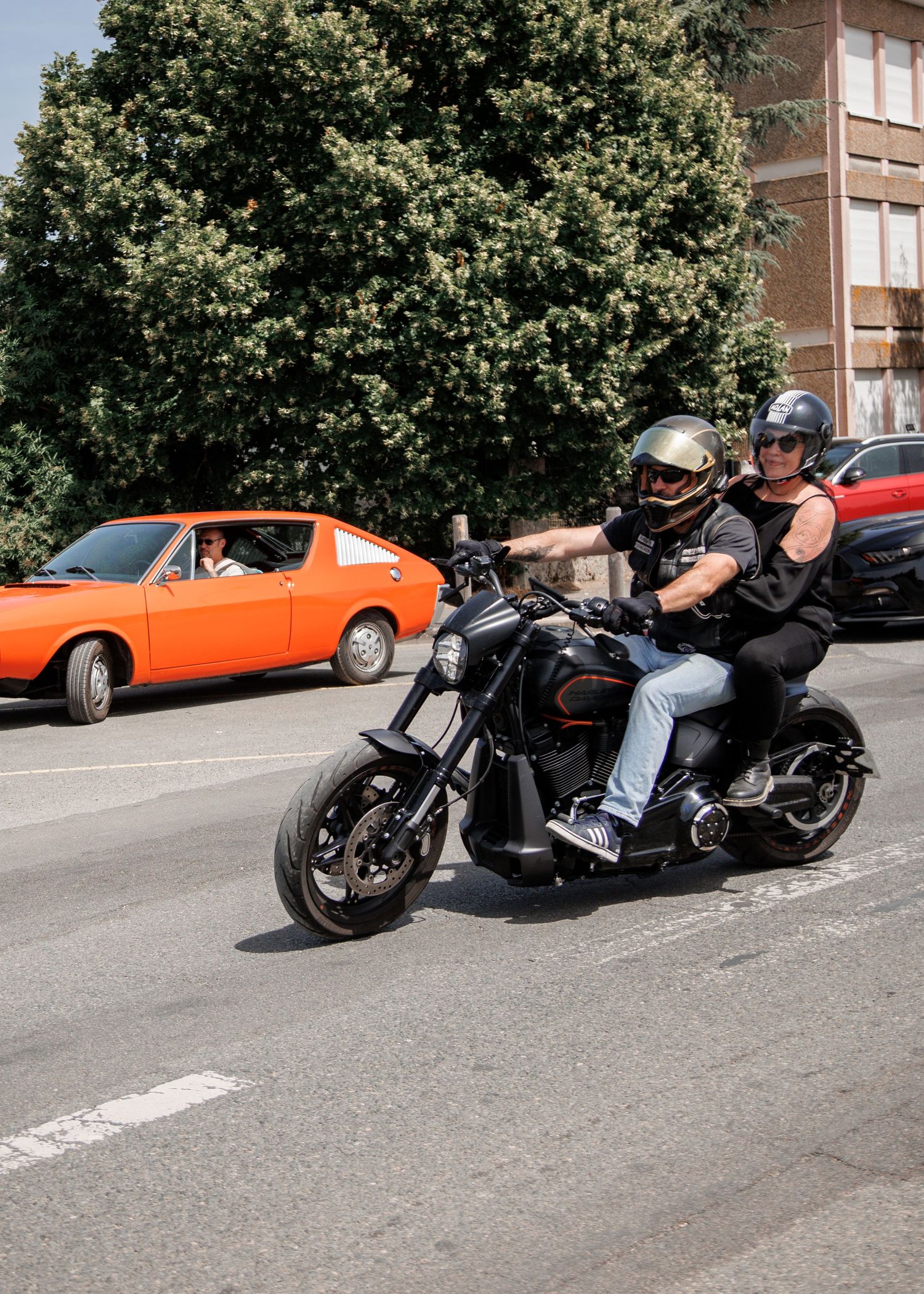 Two riders on a black cruiser motorcycle share the road with a classic orange car on a sunny day.