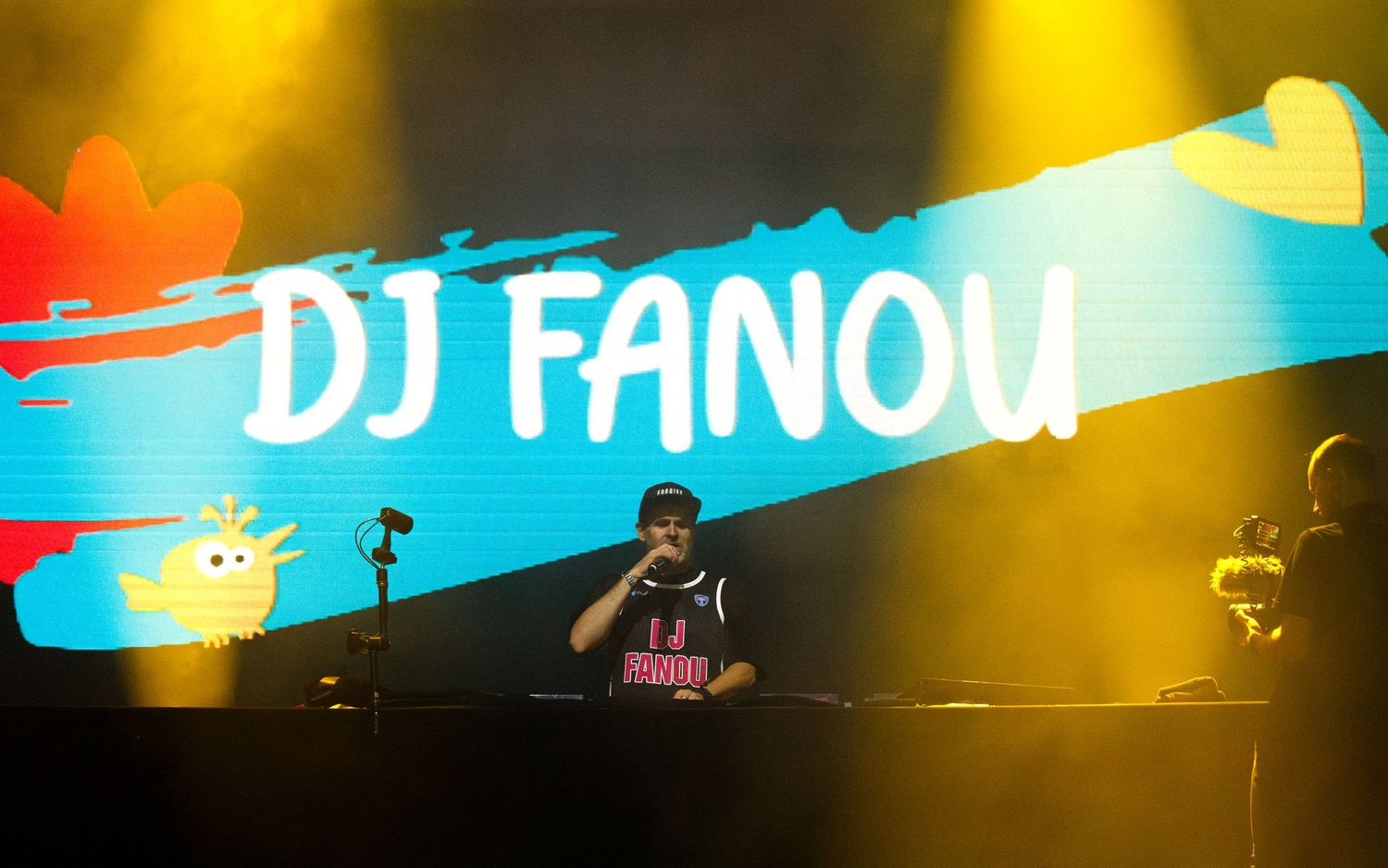 DJ performs behind decks on stage with colorful yellow lights and blue banner graphic displaying 
