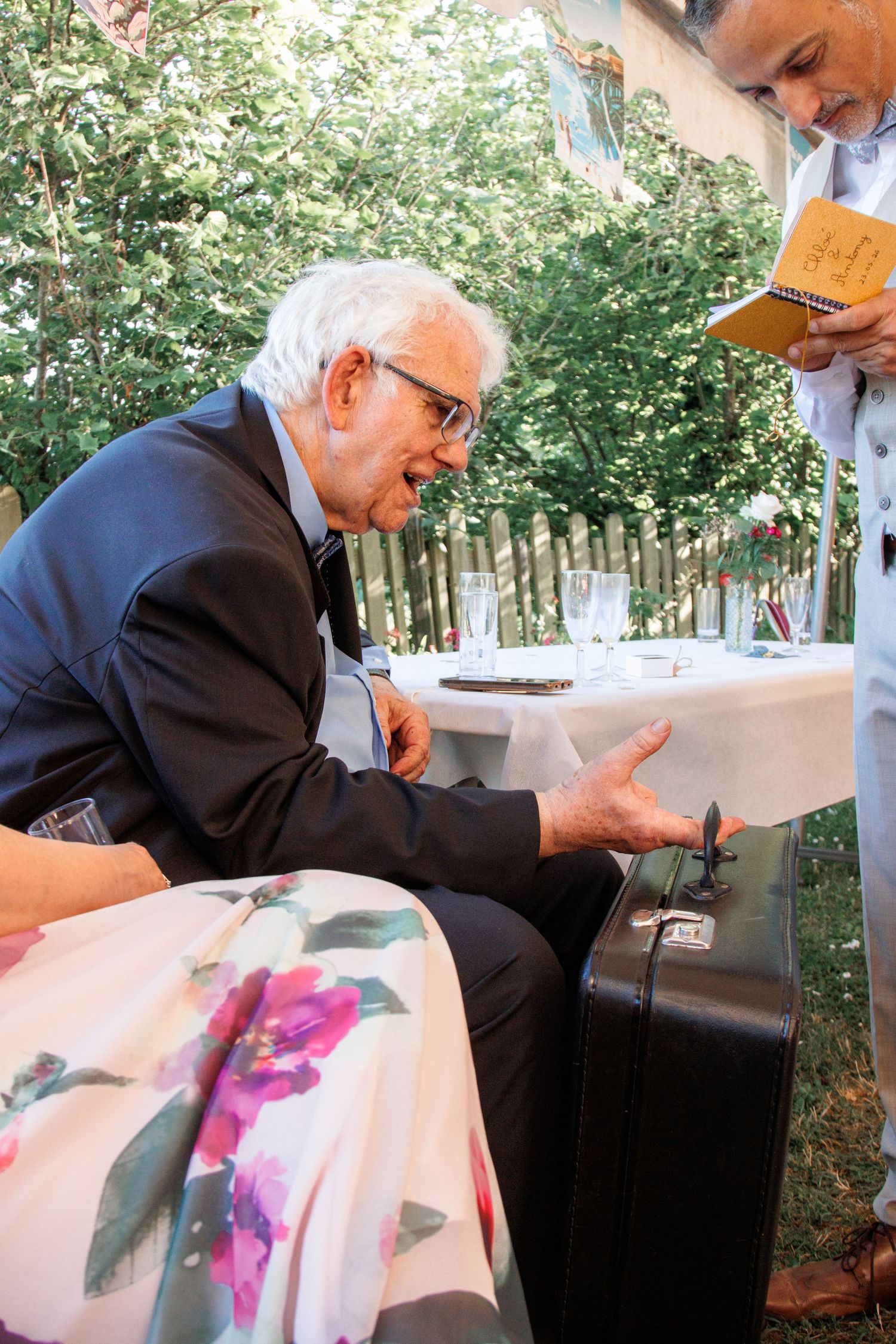 Elderly man in suit gesturing expressively while seated outdoors at wedding reception.