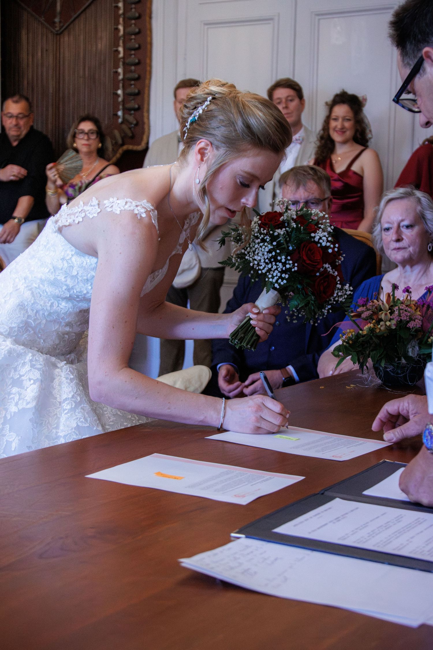 Bride in lace wedding gown signs marriage certificate at wooden table.