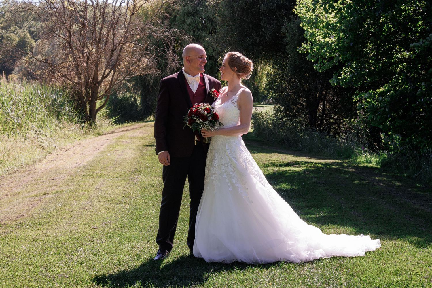 Bride and groom posing together outdoors on green lawn with trees in background on wedding day.