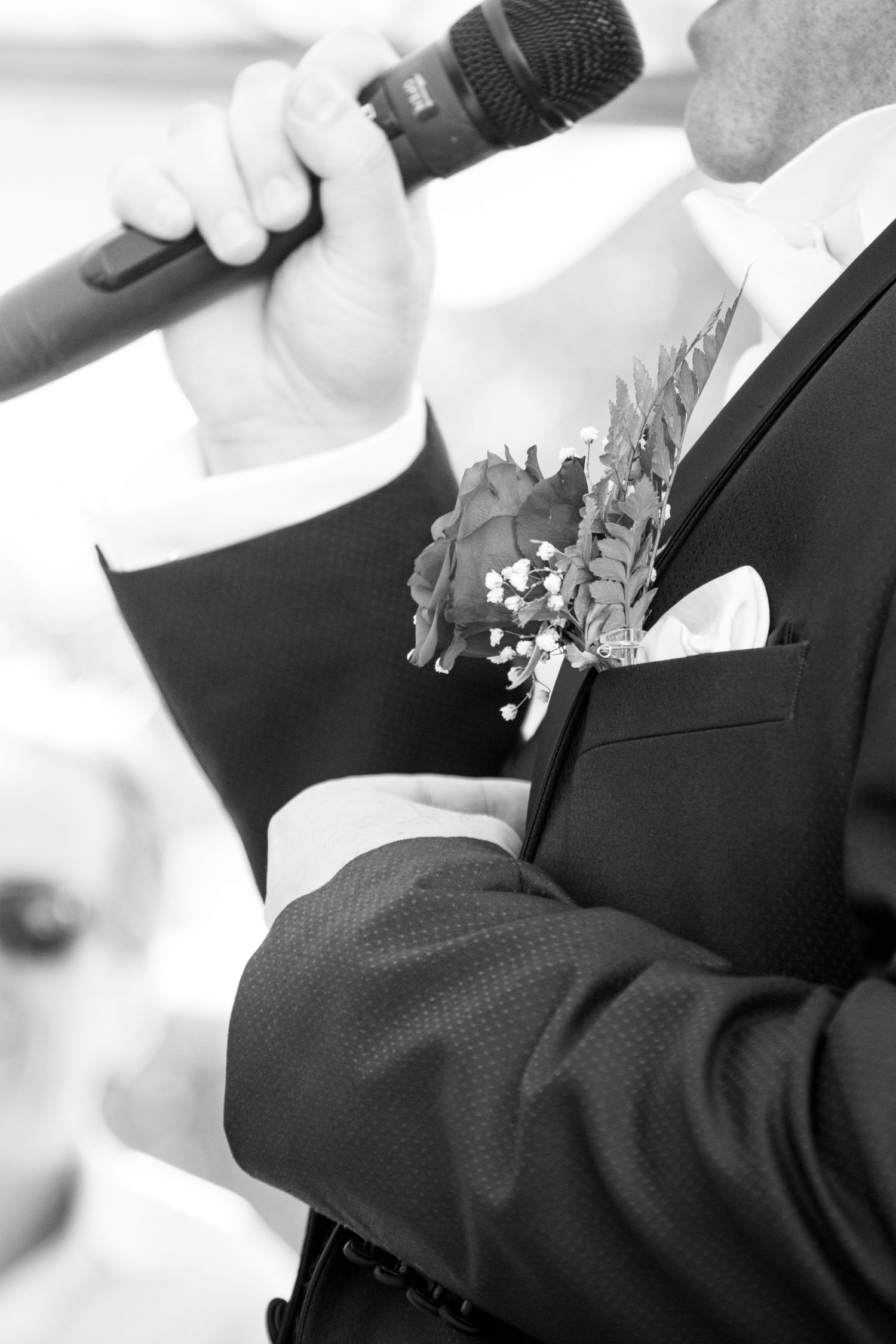 Black and white close-up of groom holding microphone with floral boutonniere on suit lapel.