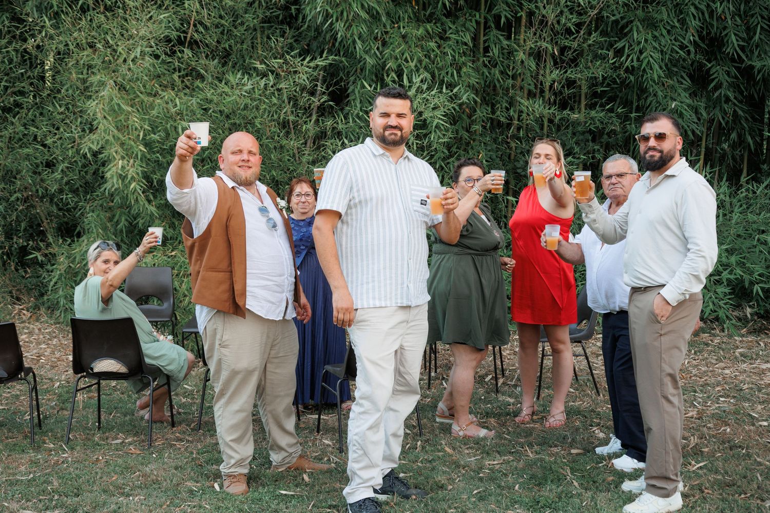 Wedding guests raising glasses in a toast outdoors, with a man in white shirt at center.