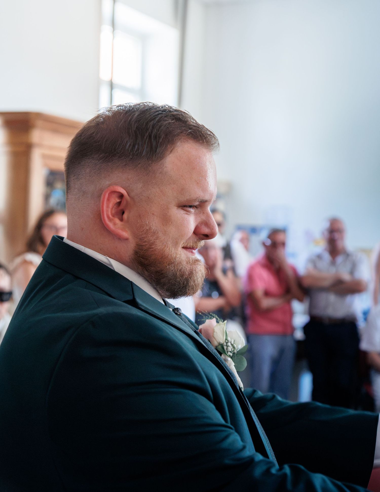 Emotional groom in dark green suit with boutonniere tearfully awaiting his bride at wedding ceremony.