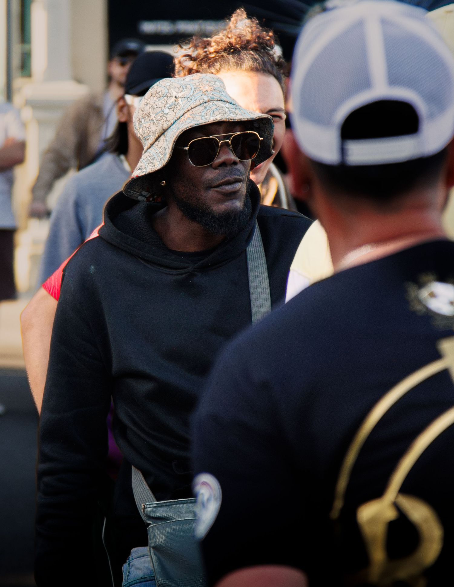 Person wearing sunglasses and bandana speaks with others at outdoor gathering.