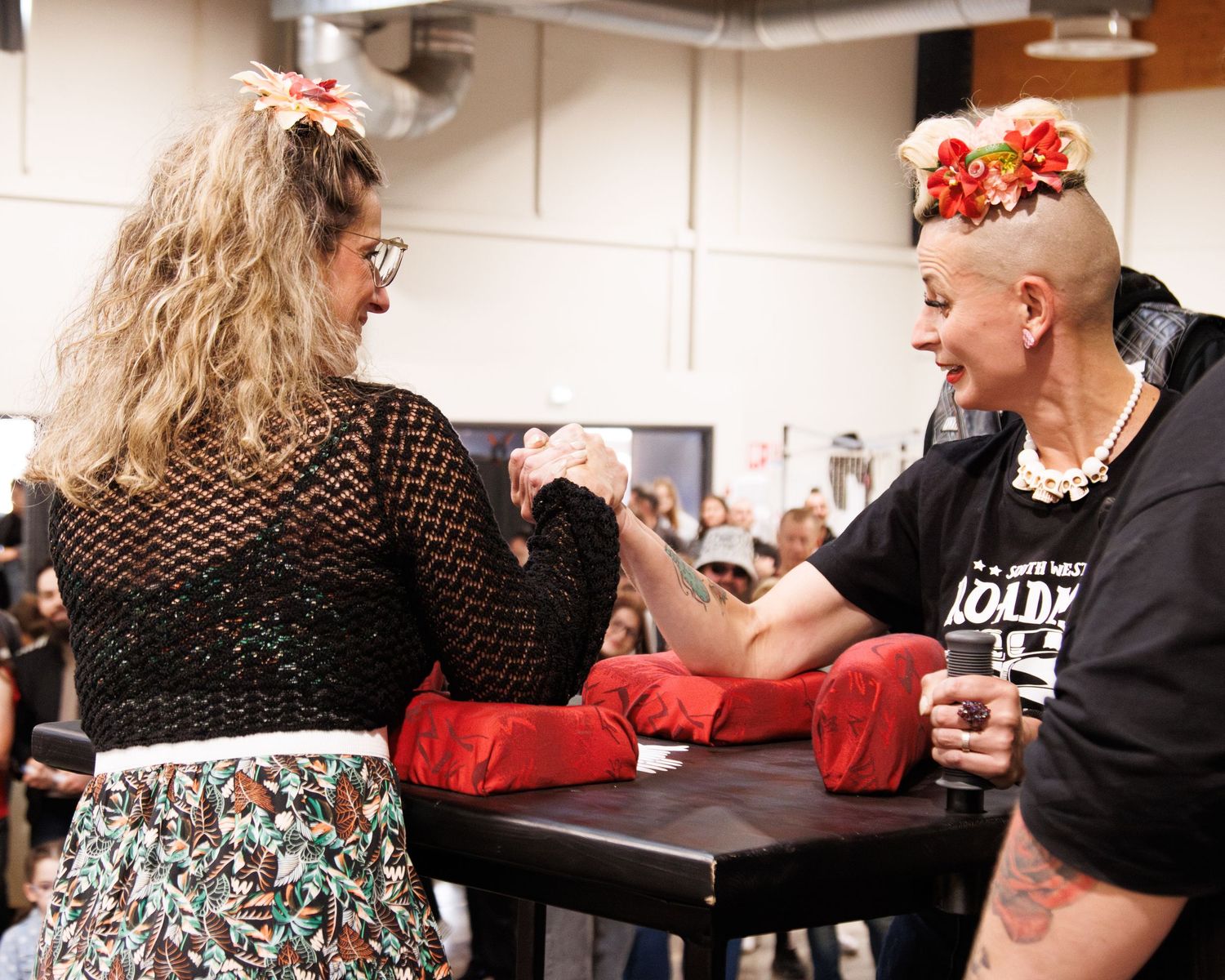 Competitive arm wrestling match on red padding with spectators watching.