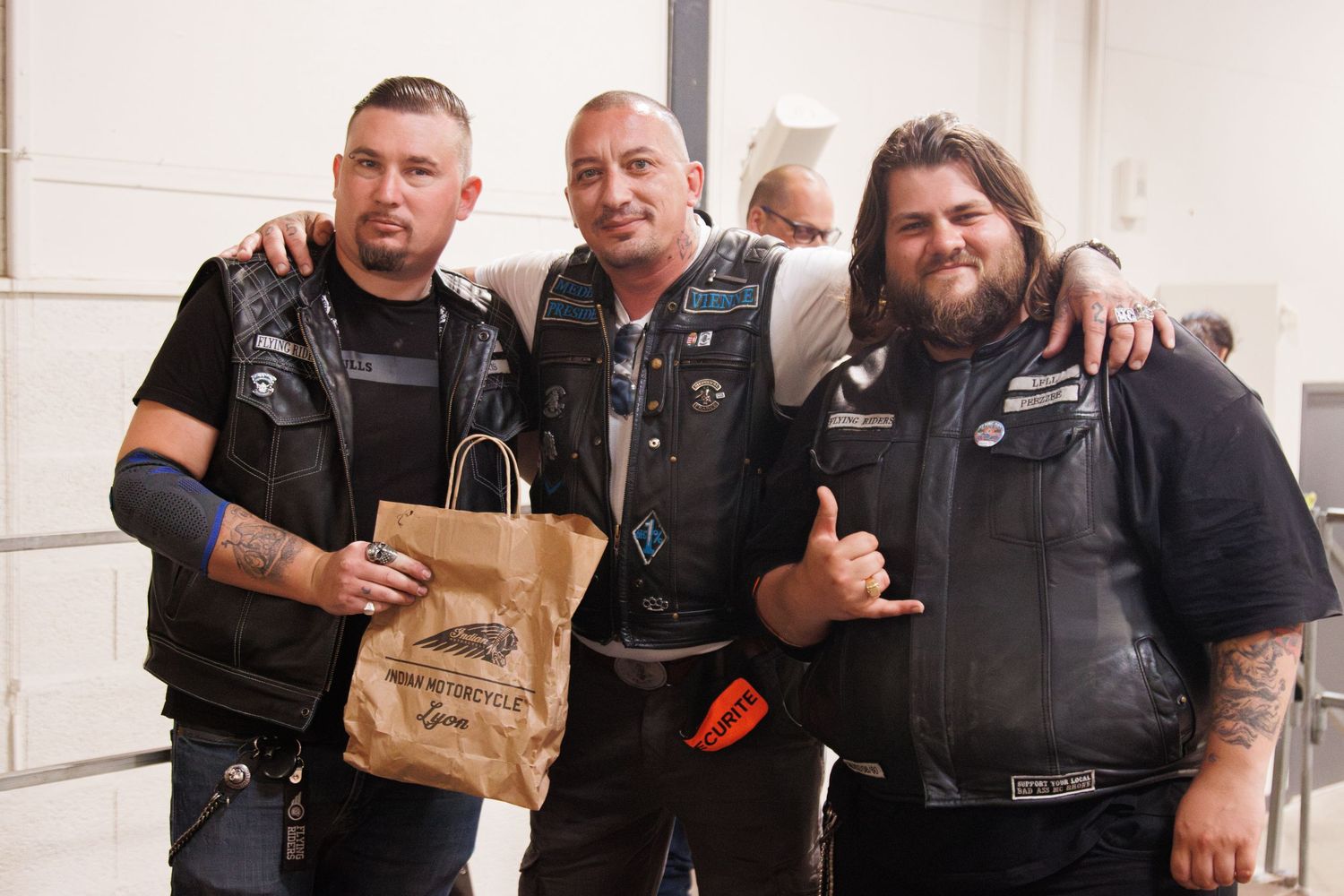 Three motorcycle enthusiasts in black leather vests smile and pose for a casual photo indoors.