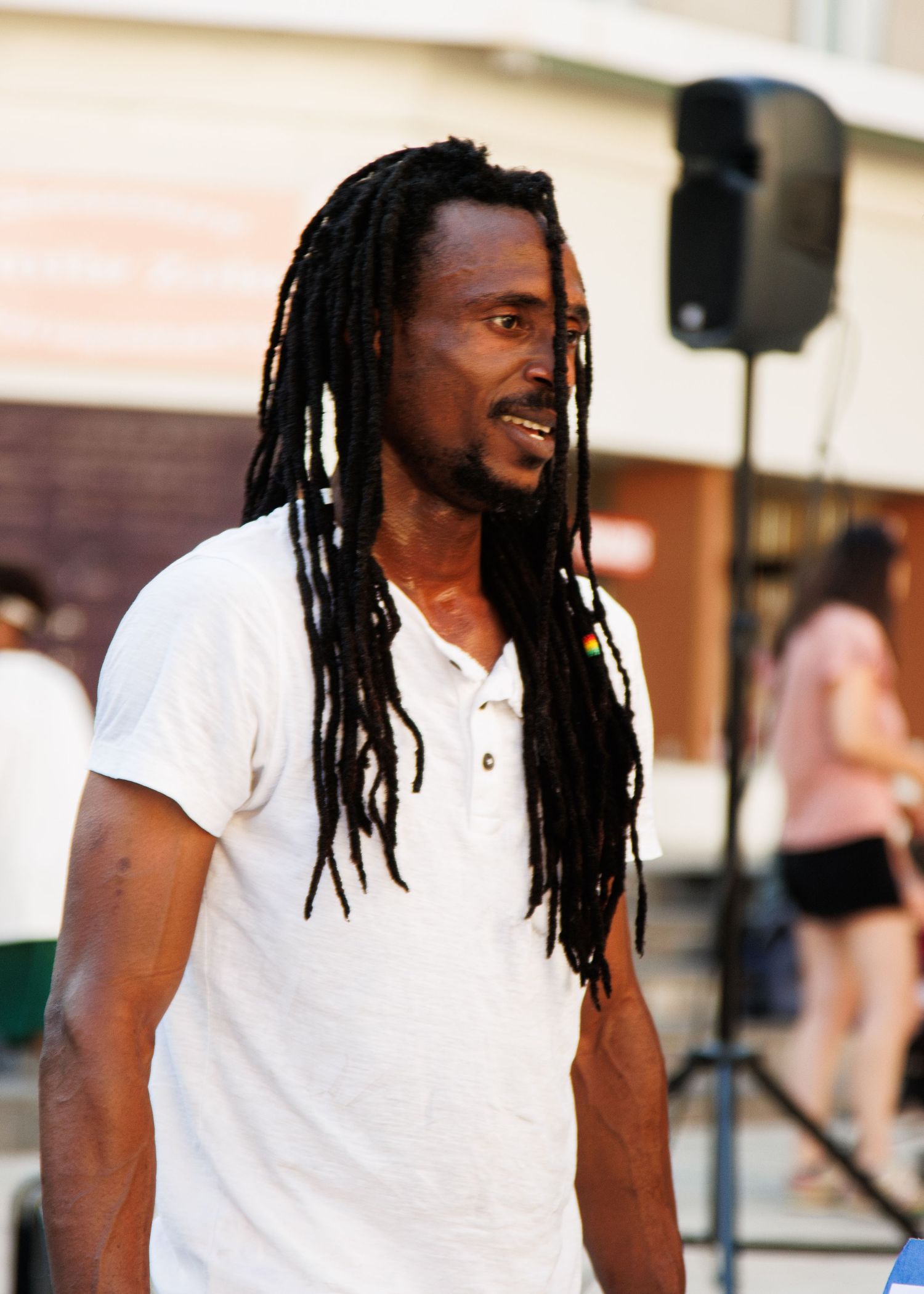 Performer with dreadlocks wearing white shirt stands before outdoor sound equipment.