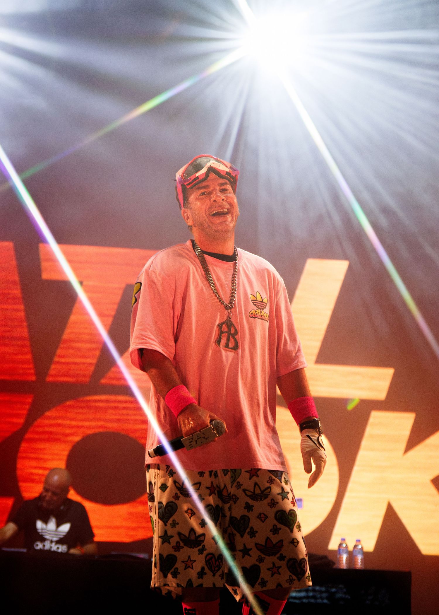 An energetic performer in a pink shirt stands on stage with dramatic lighting and colorful laser beams during a concert.