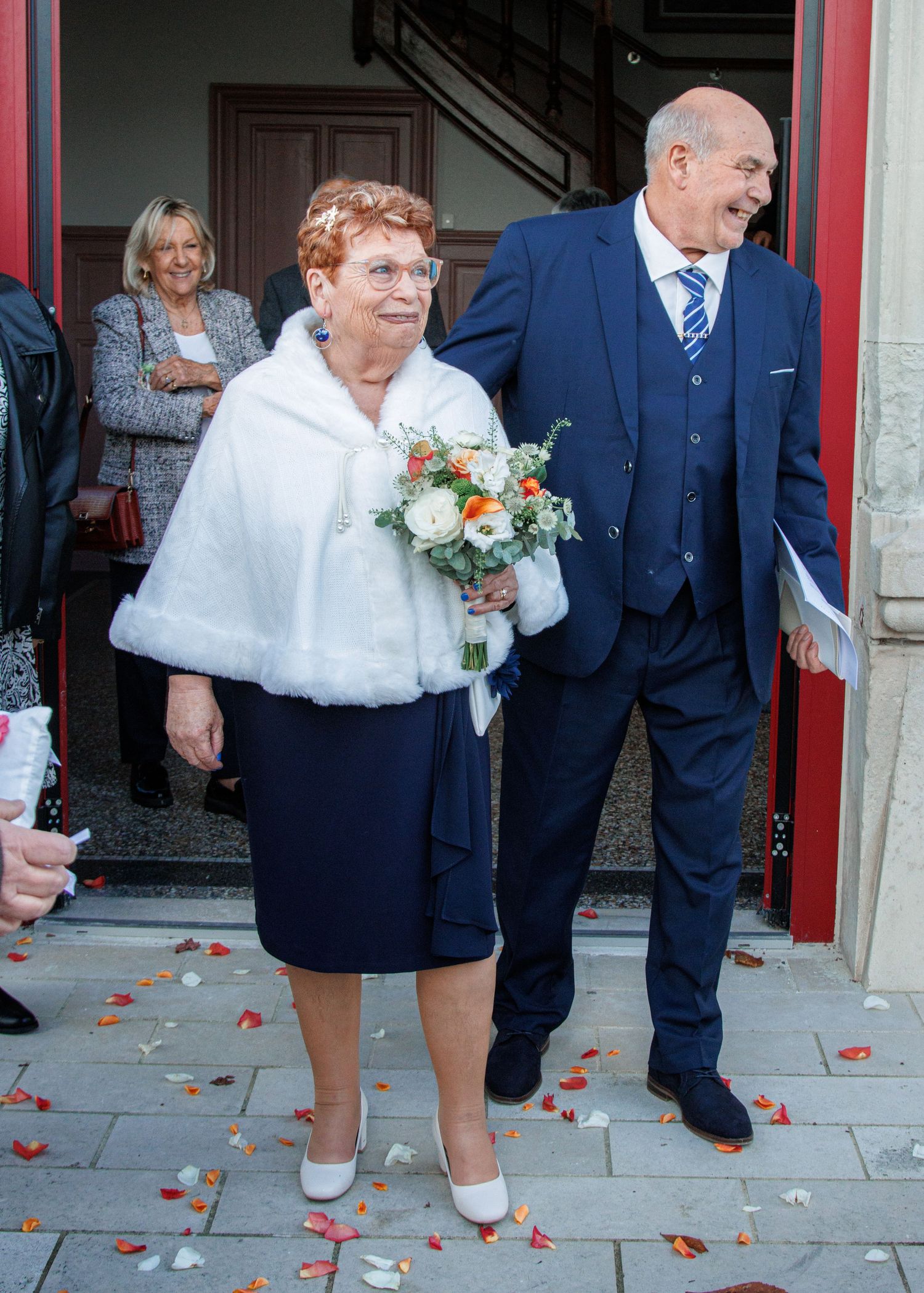 Elderly couple exits church after wedding ceremony, bride in blue dress with white fur shawl holding bouquet.