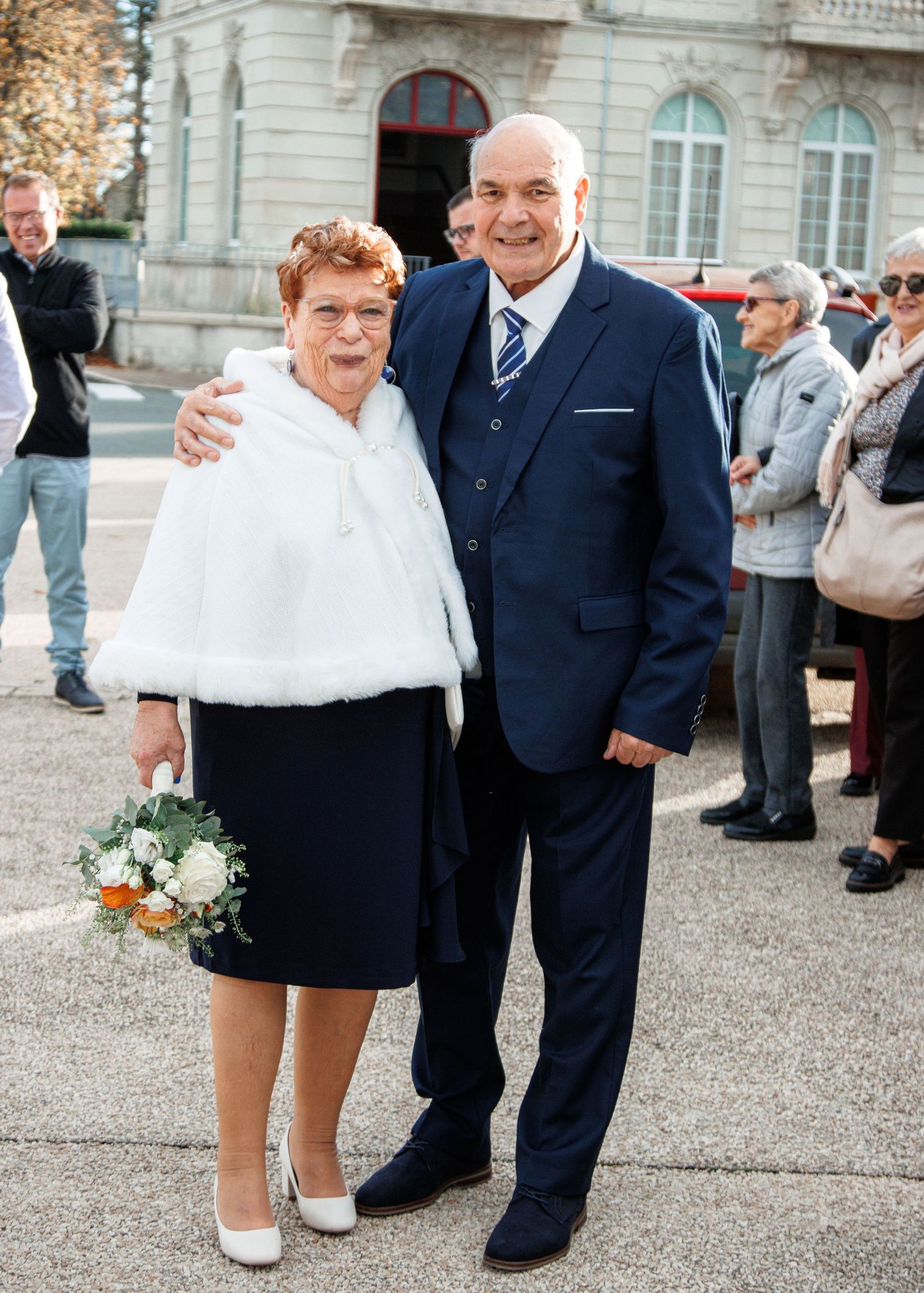 Elderly couple posing together outdoors at a wedding, woman in white fur shawl holding bouquet, man in navy suit.