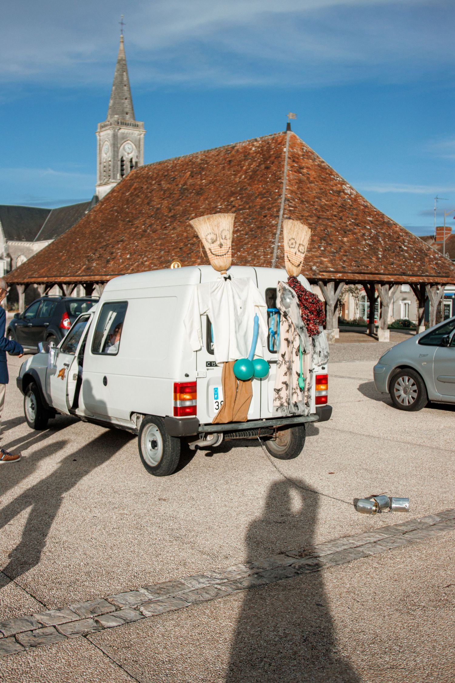 Decorated white van with puppet figures parked in a French village square near a historic church and market hall.