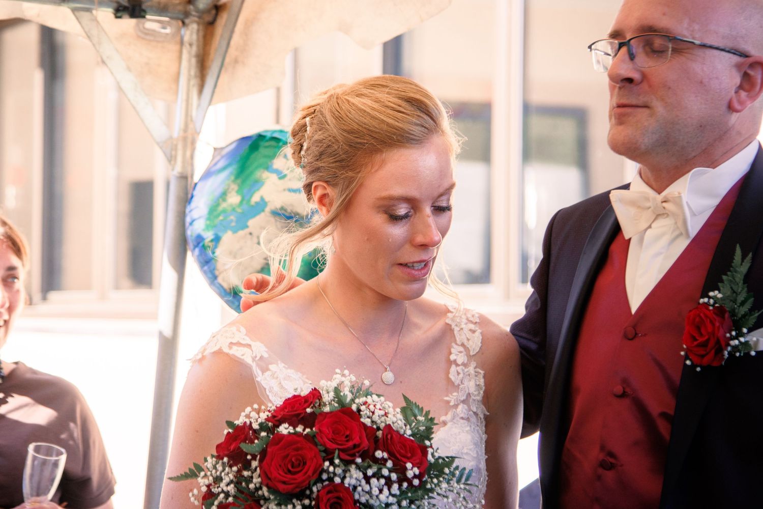 Close-up of bride holding red rose bouquet with groom in burgundy vest beside her at wedding ceremony.