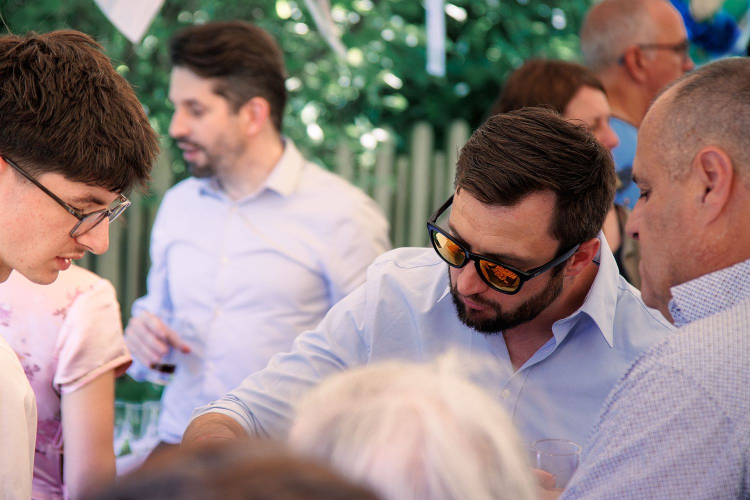 Bearded man in sunglasses leans over table chatting with guests at outdoor wedding reception.