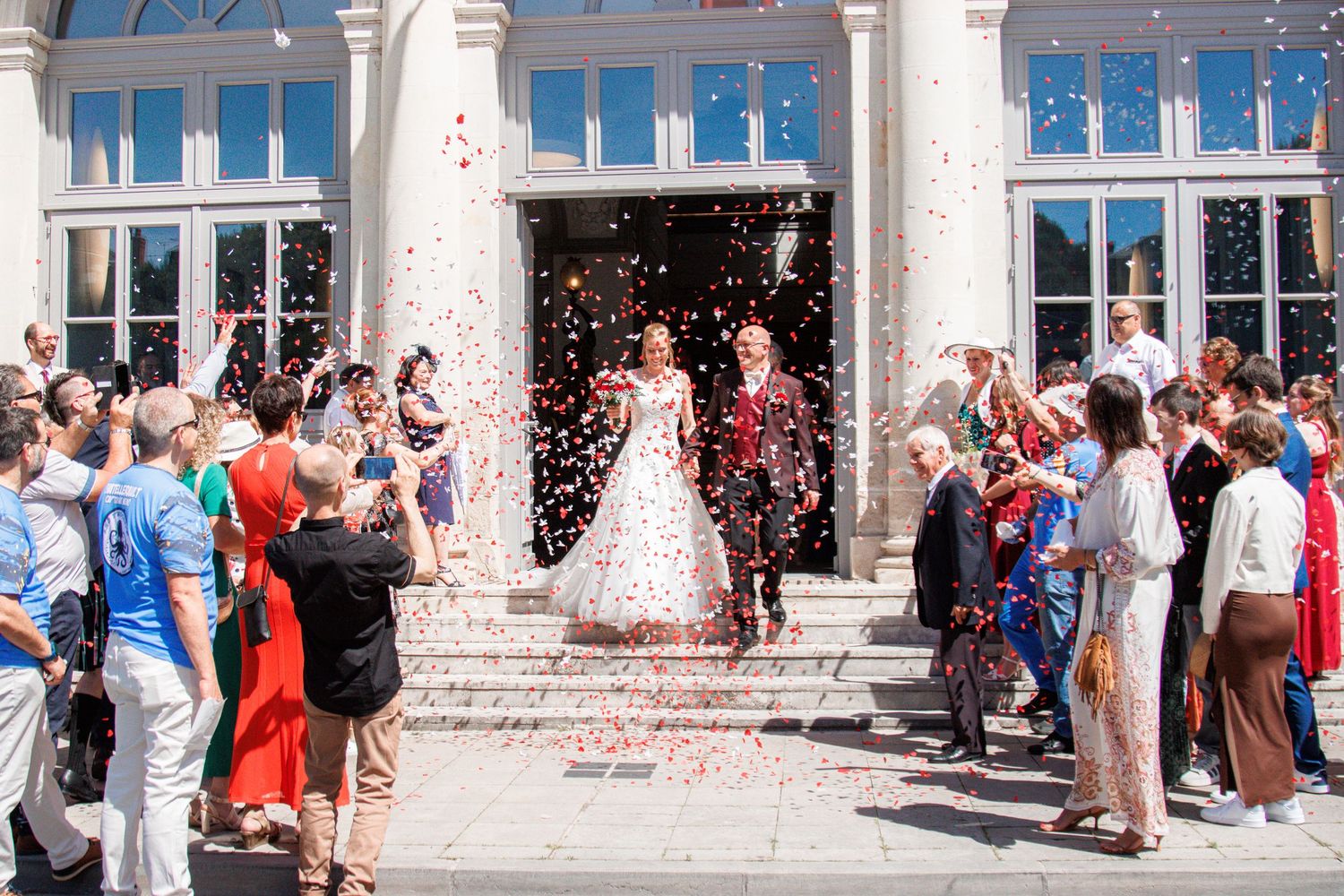 Joyful newlyweds exit ceremony venue as guests shower them with colorful confetti in sunny outdoor setting.