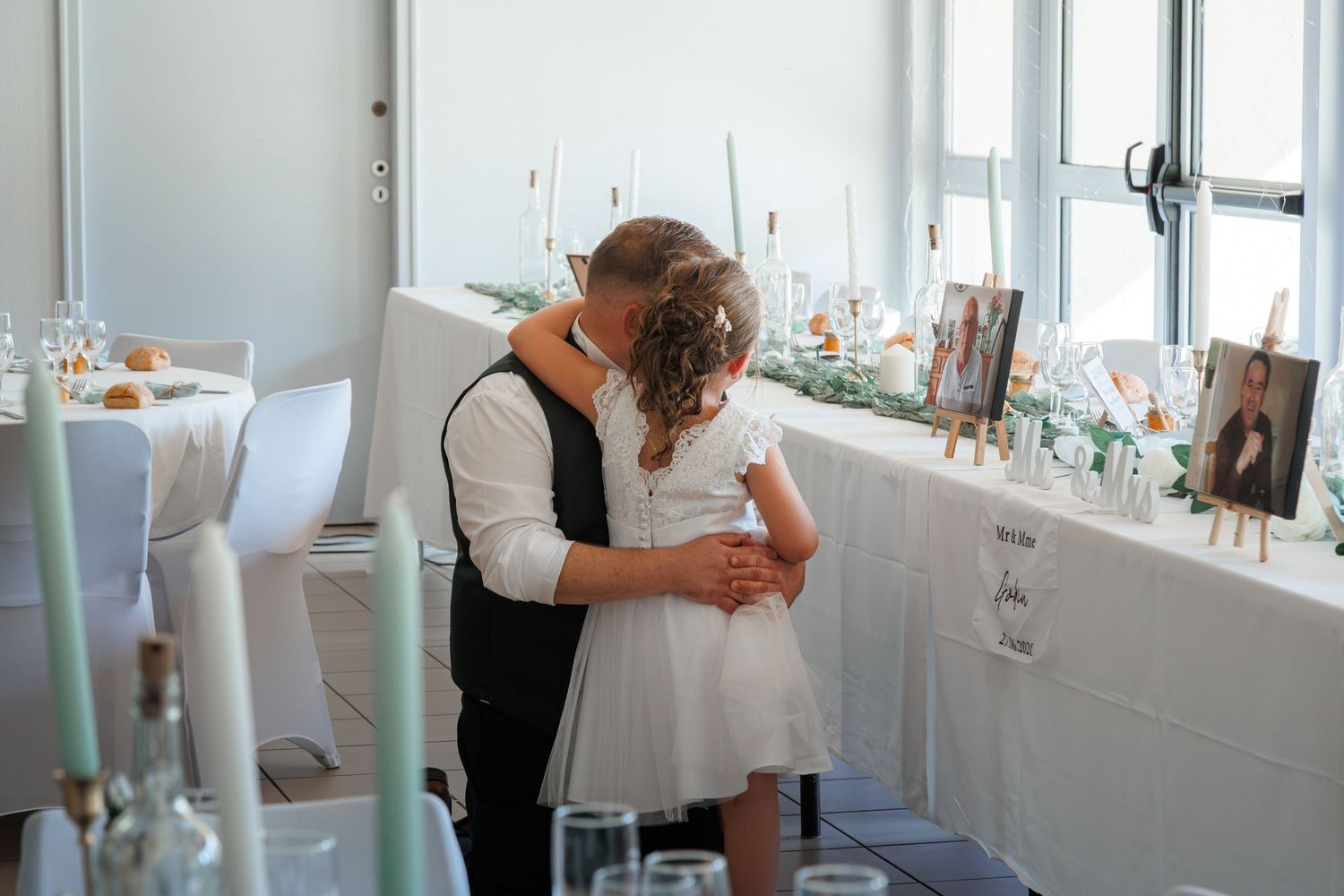 Bride and groom share a tender embrace at elegantly decorated wedding reception with white tablecloths and floral centerpieces.
