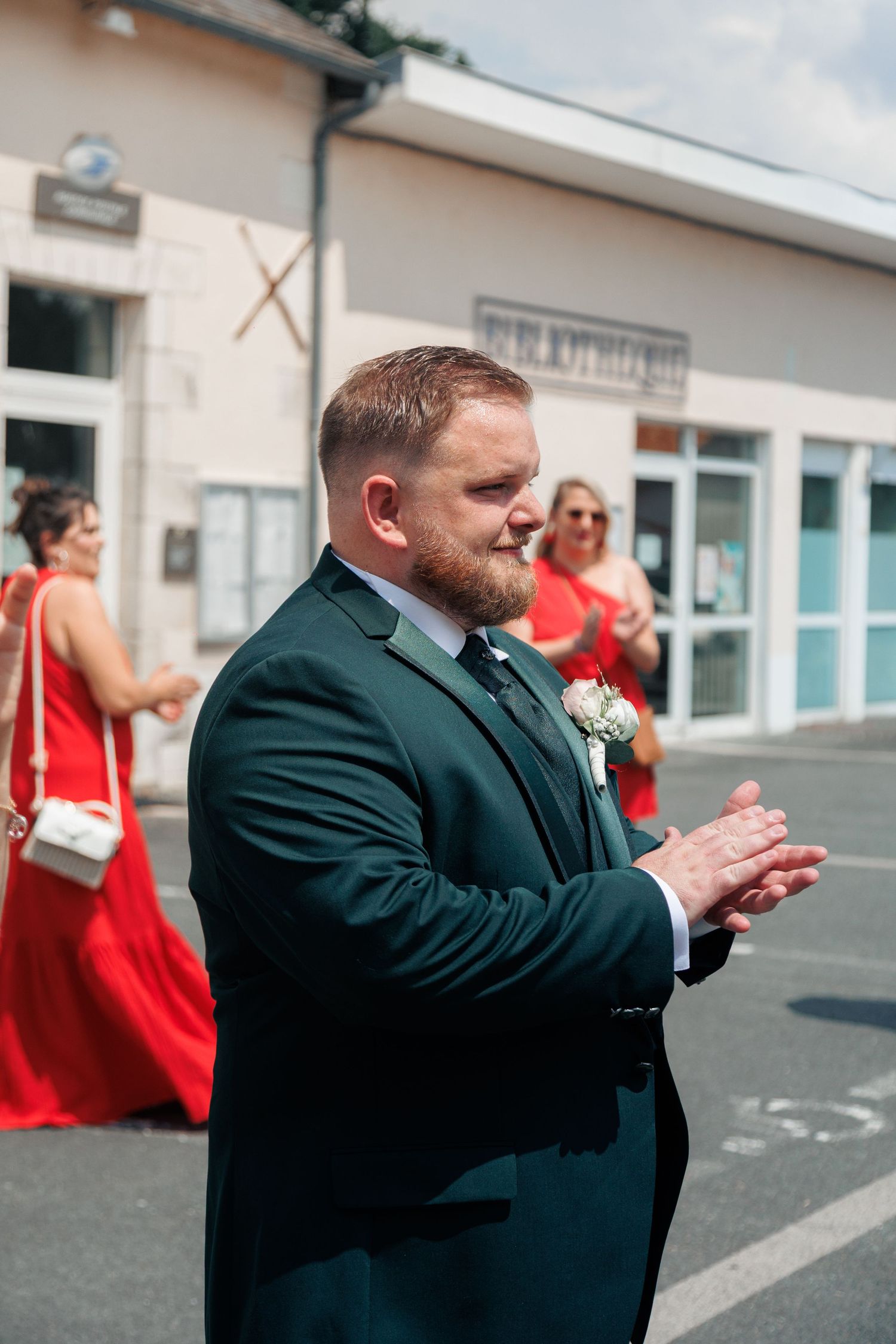 Groom in dark green suit clapping outdoors at wedding with bridesmaids in red dresses in background.