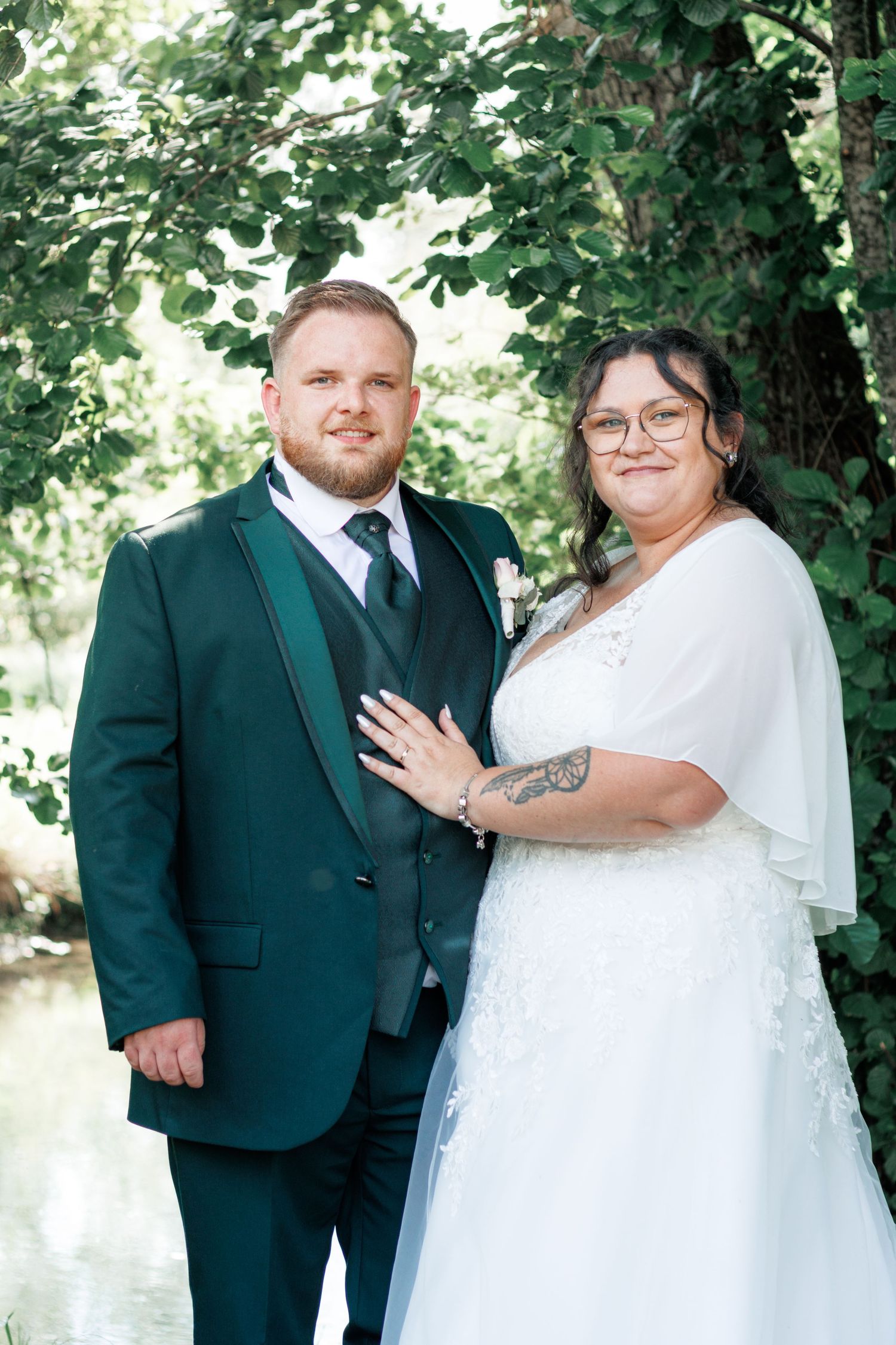 Bride and groom posing together in green suit and white wedding dress under lush foliage.