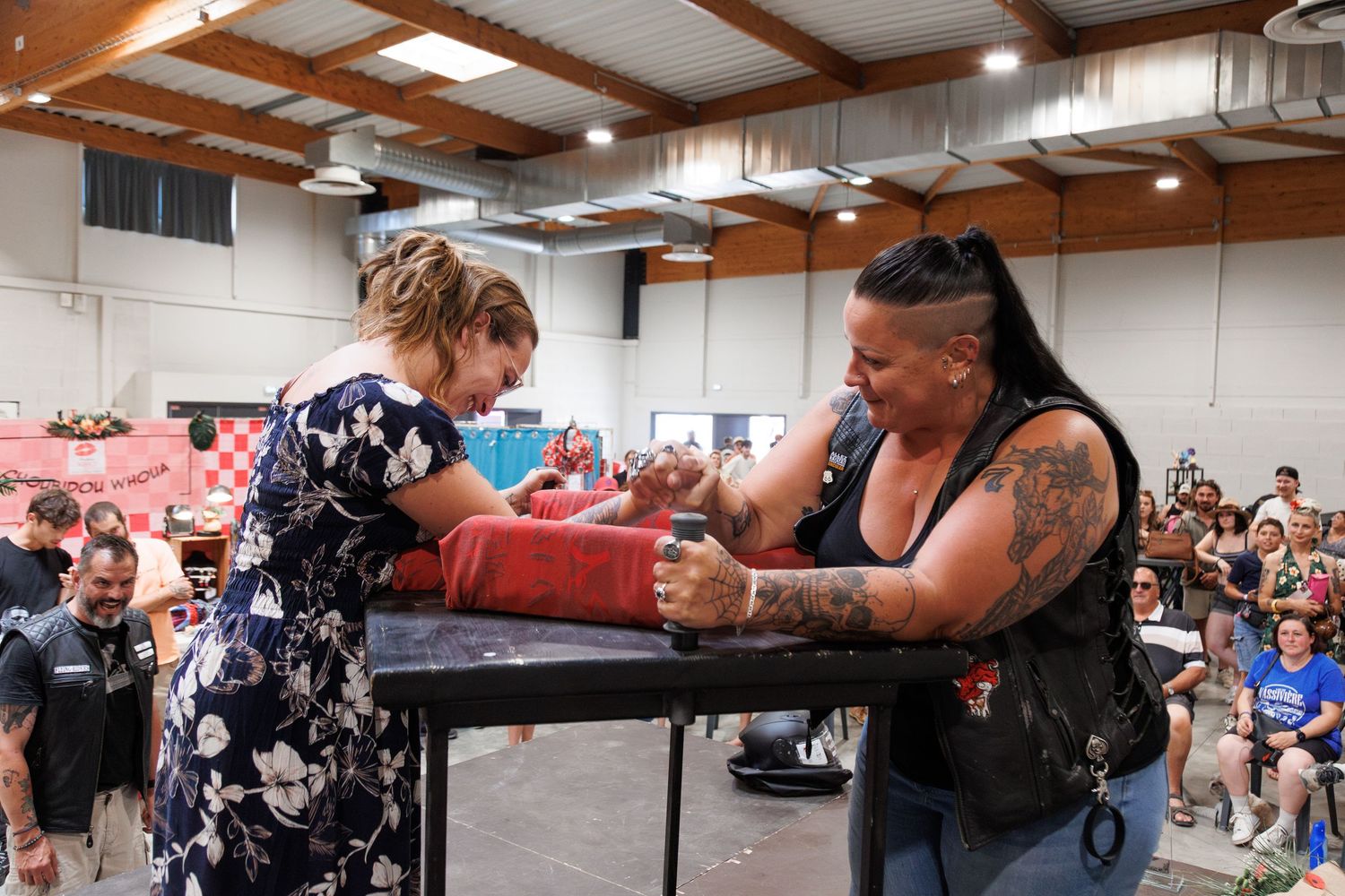 Two women arm wrestling at a table during a competitive event inside a large indoor venue with spectators.