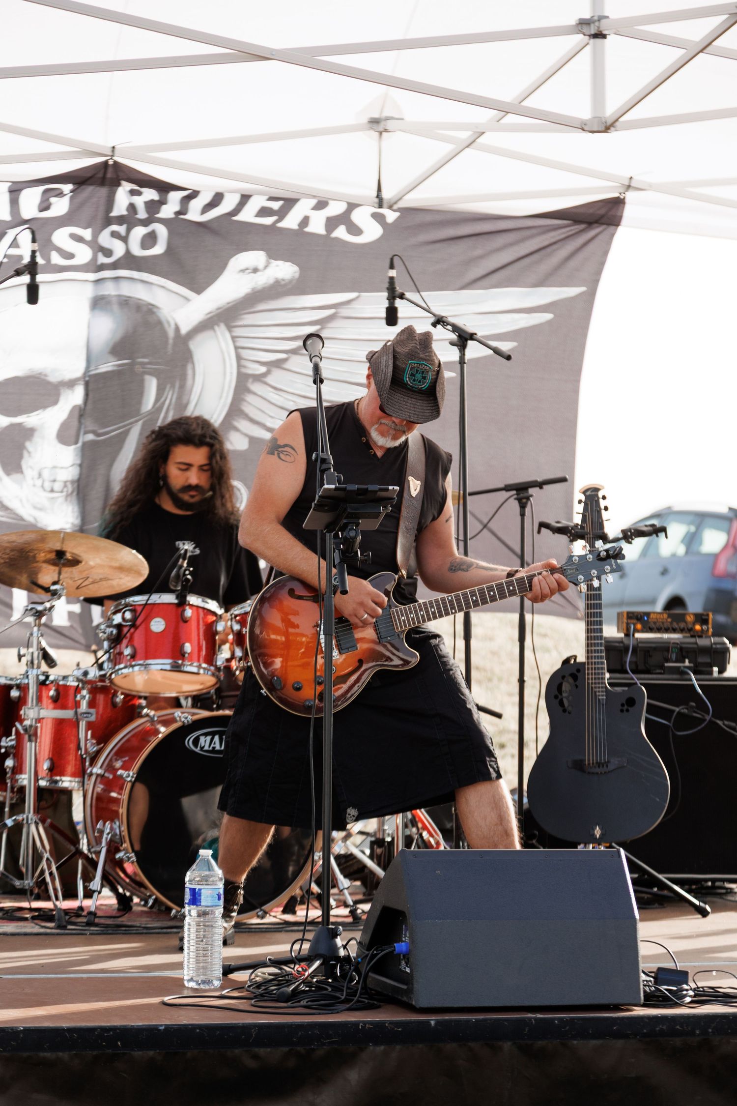 Rock band performing outdoors at a biker rally, guitarist playing electric guitar on stage with drummer behind red kit.