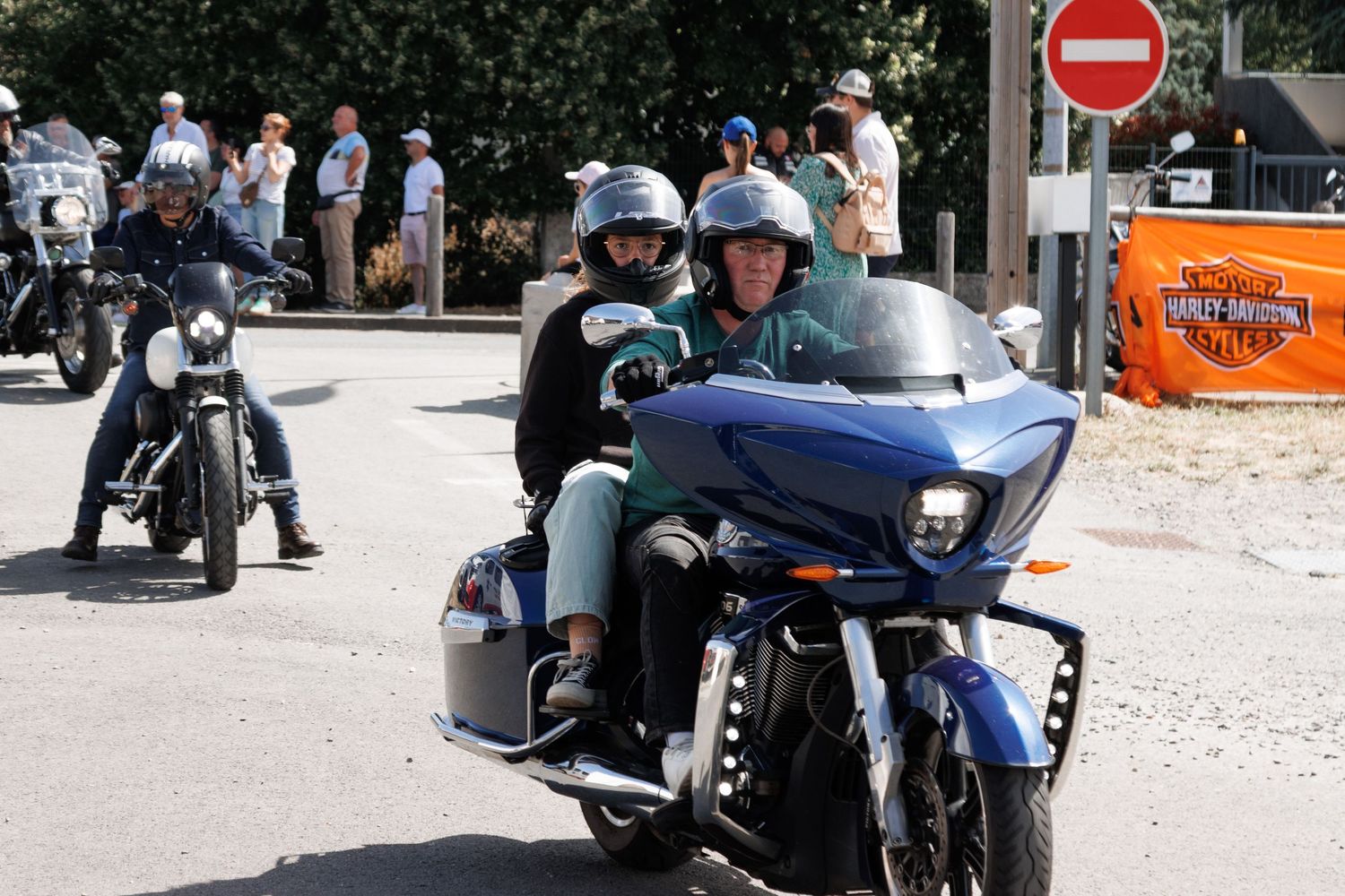Motorcyclists riding blue and black bikes past a no-entry sign at a Harley-Davidson event on a sunny day.