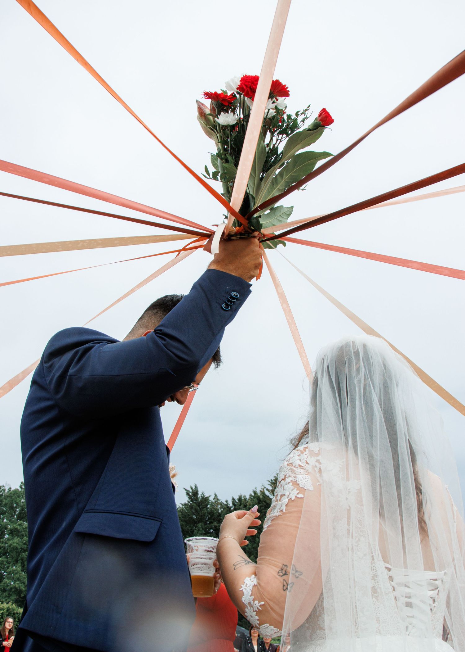 Bride and groom holding red rose bouquet with colorful ribbons at outdoor wedding ceremony.