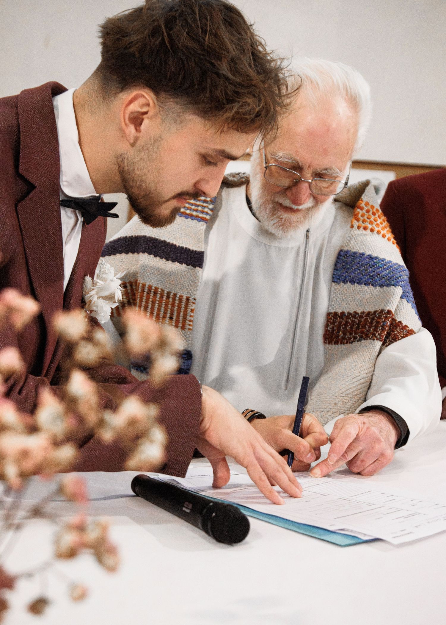 Young groom signing wedding documents with elderly officiant in white robe and colorful vest at ceremony table.