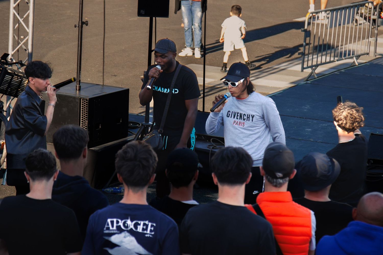 Performers on stage speaking to audience during outdoor event with speakers and lighting setup.