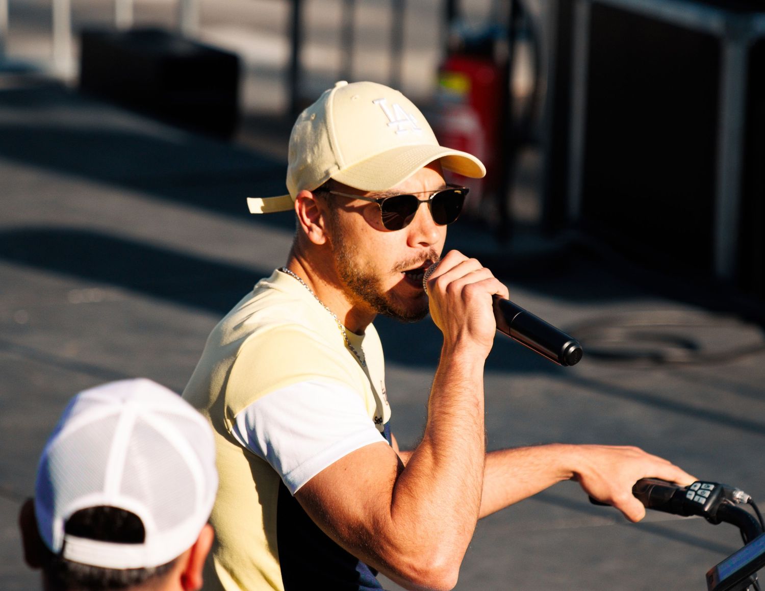 Singer wearing sunglasses and baseball cap performs on outdoor stage in summer.