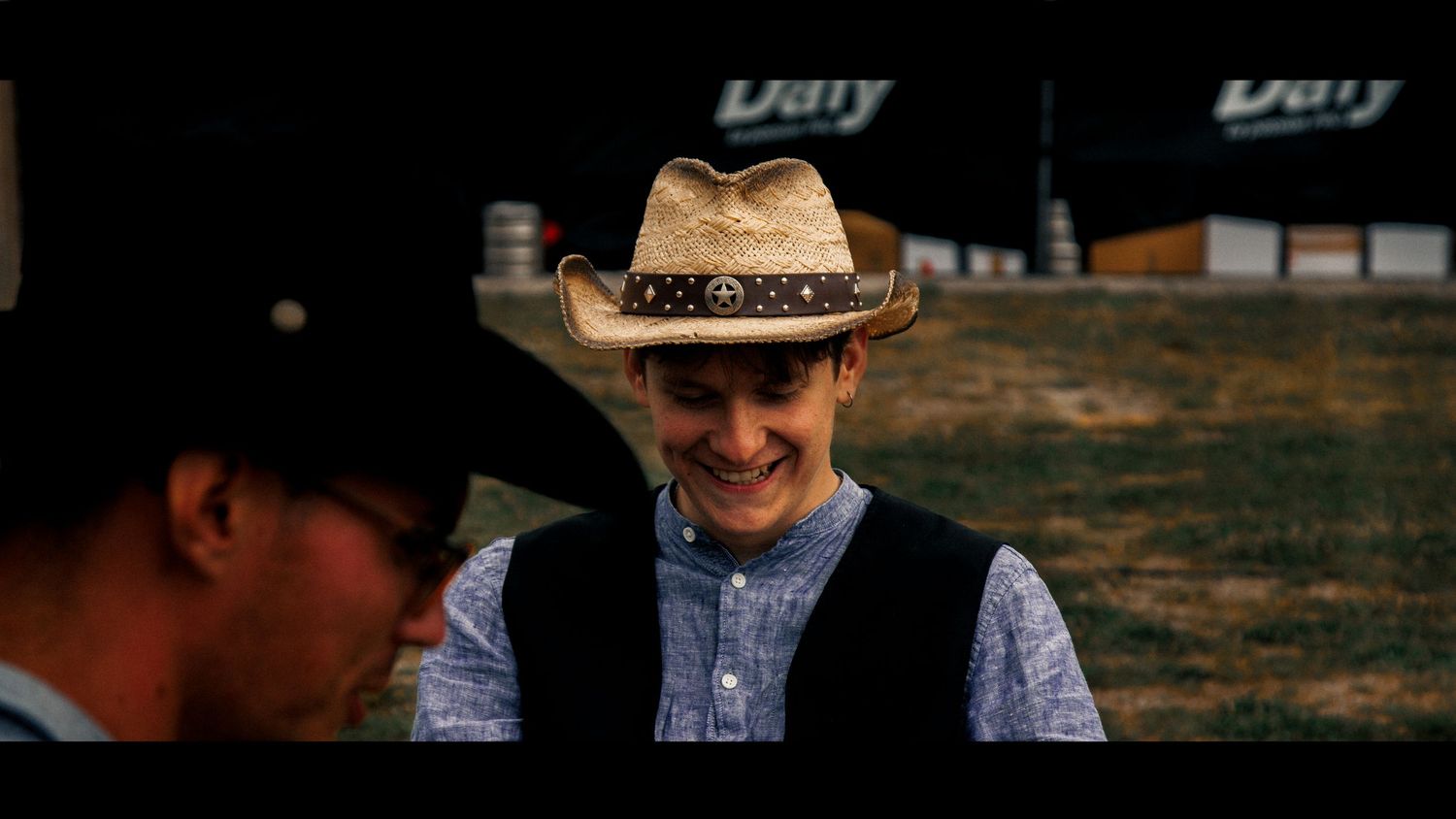 Person in western attire and cowboy hat smiles during an outdoor scene.