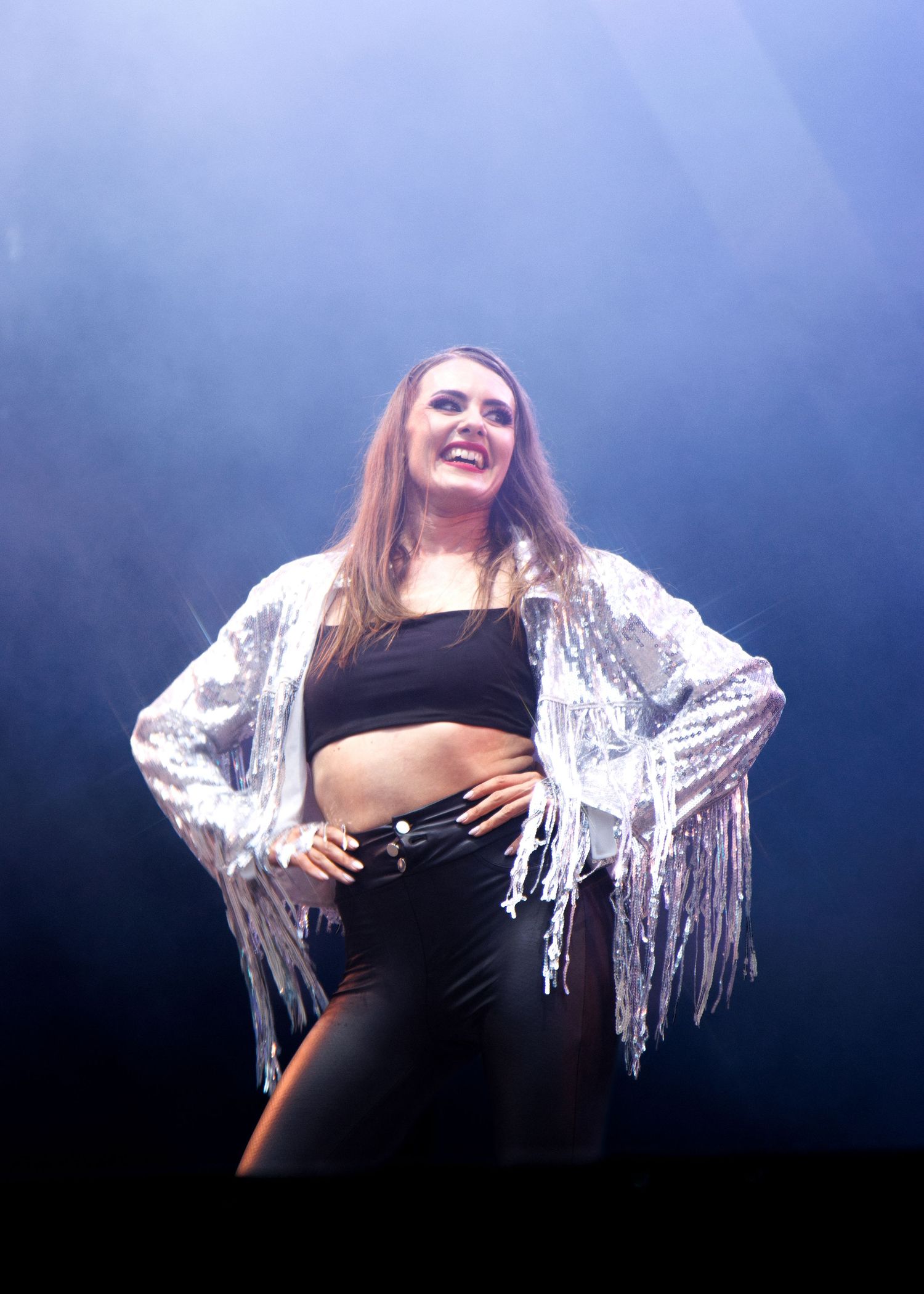 Performer dances energetically on stage wearing a sparkly silver fringe jacket and black crop top in atmospheric lighting.