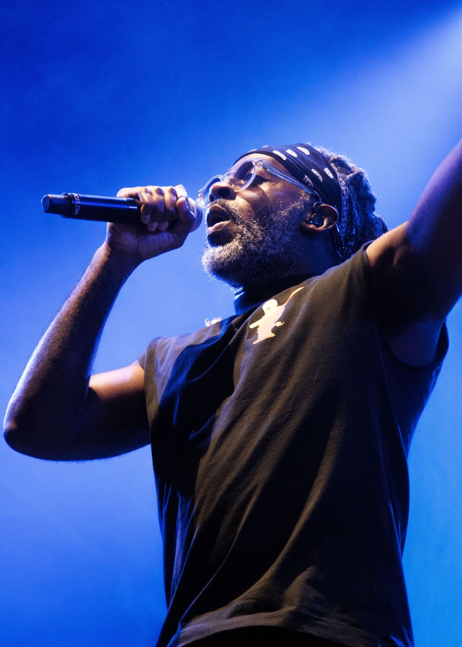 A performer in a black t-shirt sings passionately on stage with dramatic blue lighting in the background.
