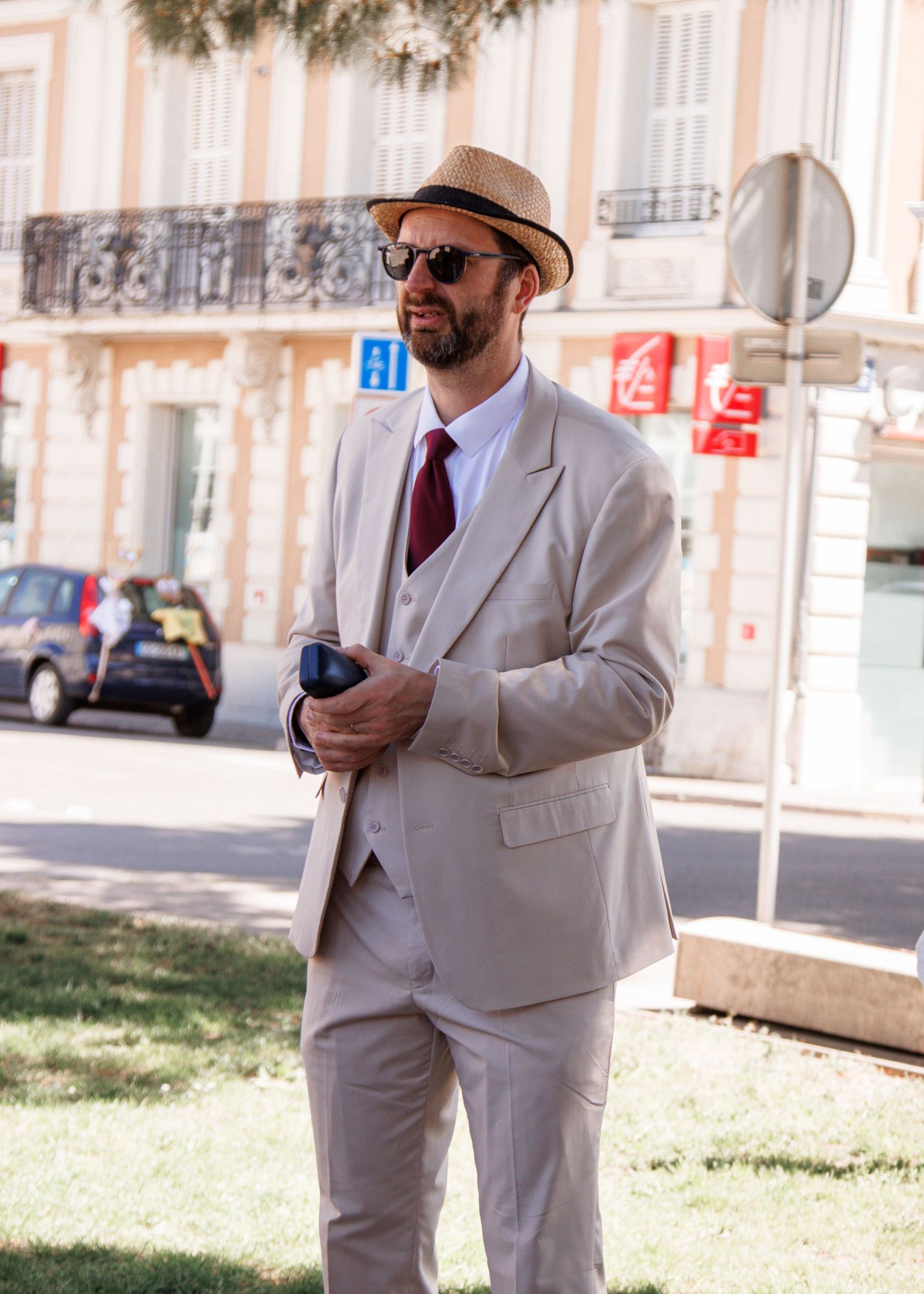 Stylish man in beige three-piece suit, red tie, straw fedora, and sunglasses standing outdoors on a sunny day.