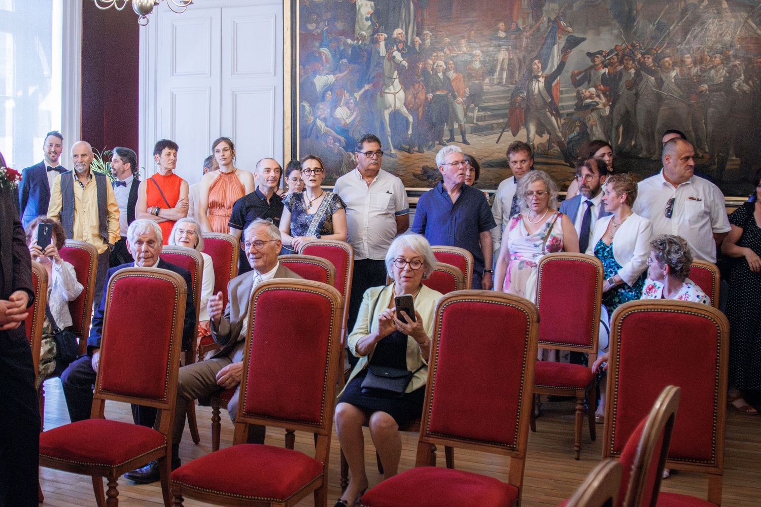 Guests gathered in an elegant hall with red chairs and a large classical painting during a formal event.