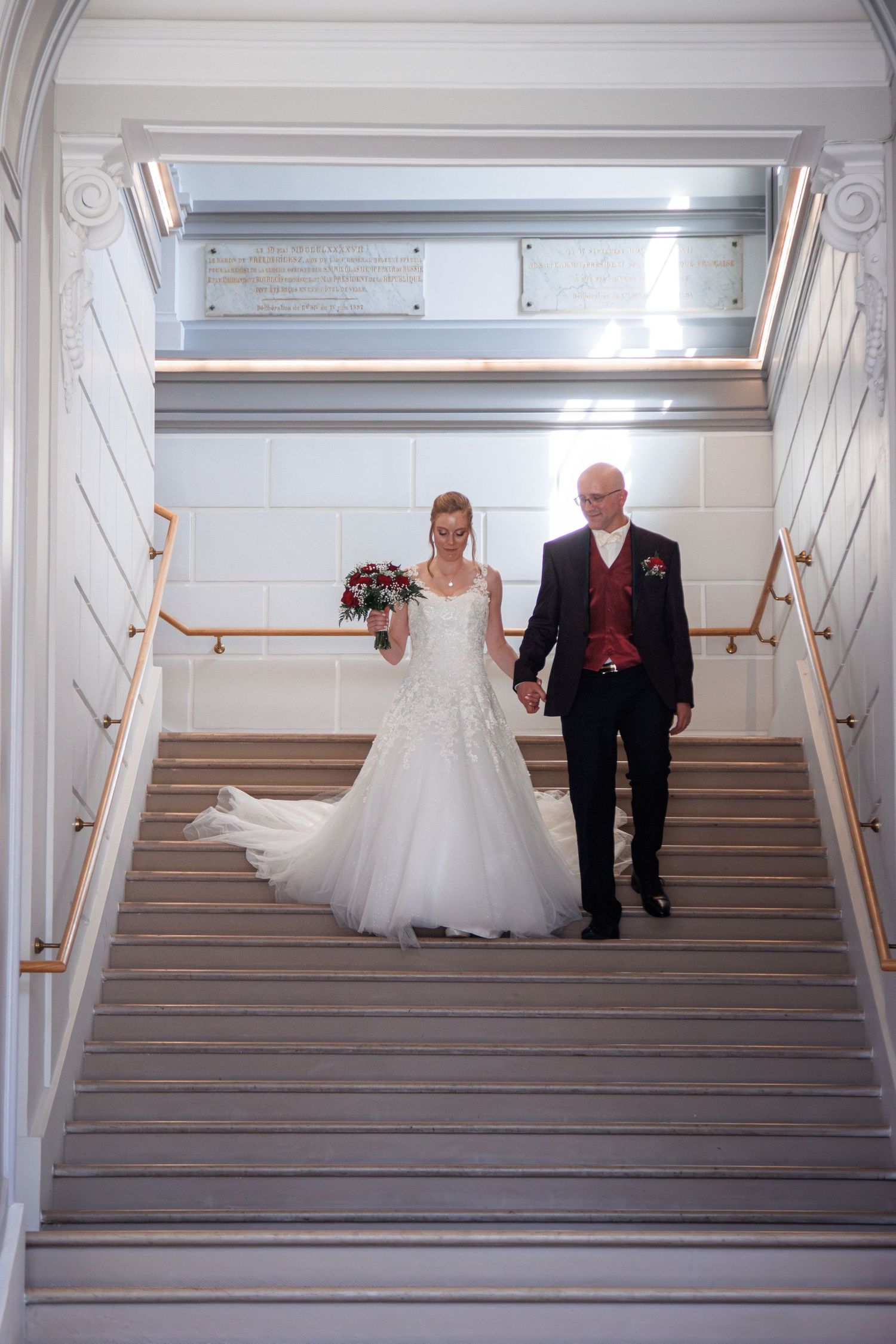 Bride and groom walking hand in hand down an elegant staircase, bride wearing a white ball gown holding a bouquet.