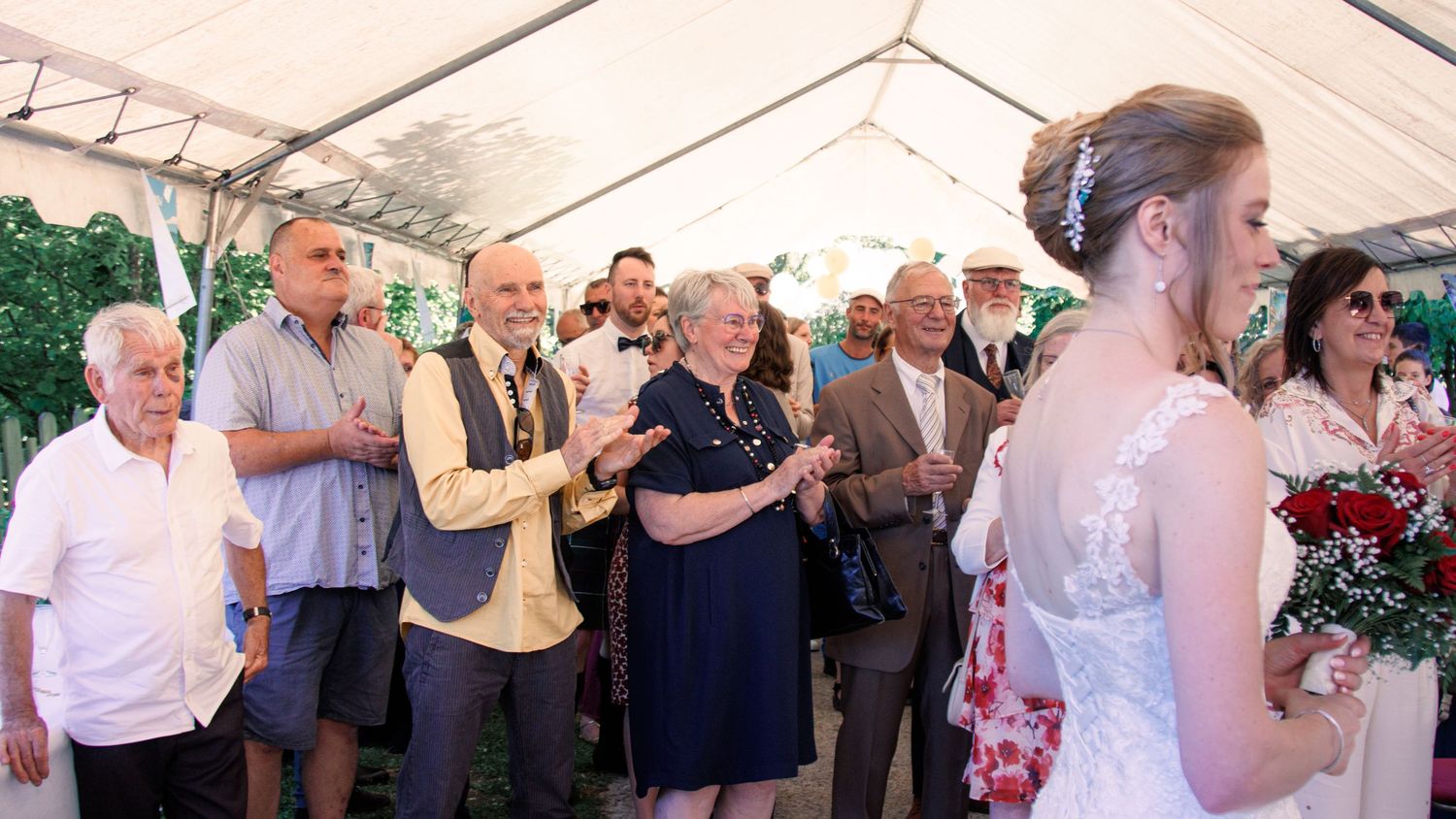 Wedding guests clapping and smiling as bride in white gown holds red roses under a tent.