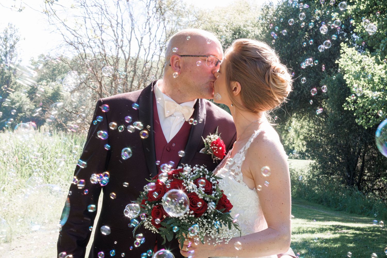 Newlyweds share a kiss surrounded by floating soap bubbles outdoors, bride holding red rose bouquet.