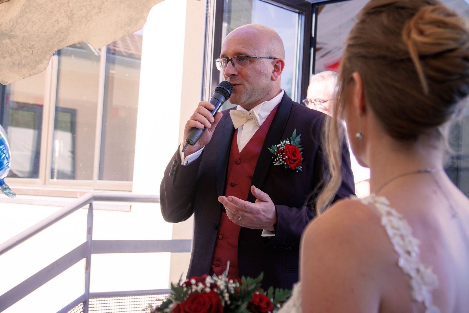 Bald groom in navy suit speaks into microphone addressing bride during outdoor wedding reception ceremony.