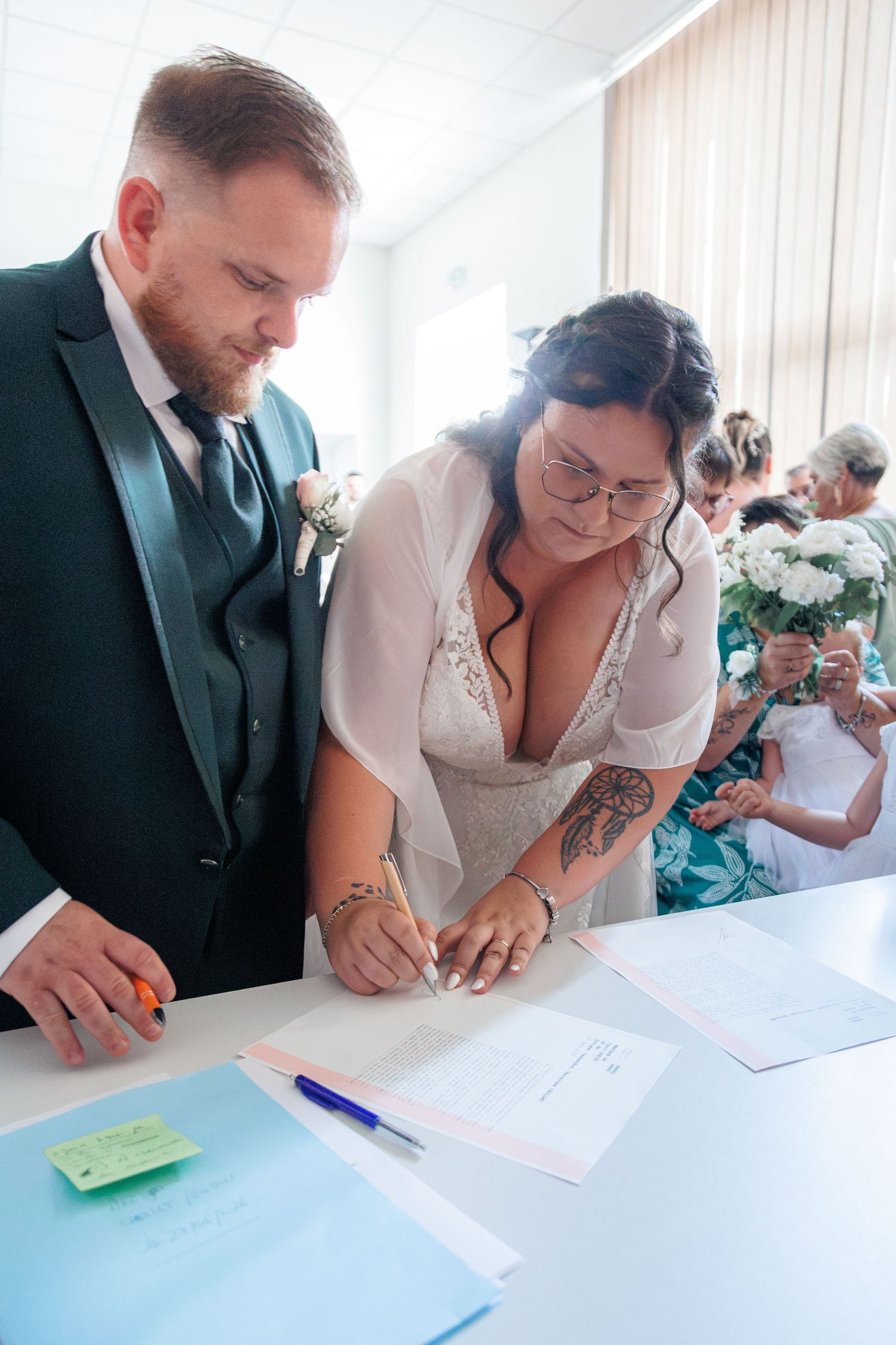 Bride with tattoos and groom in green suit sign wedding documents at a white table during their ceremony.