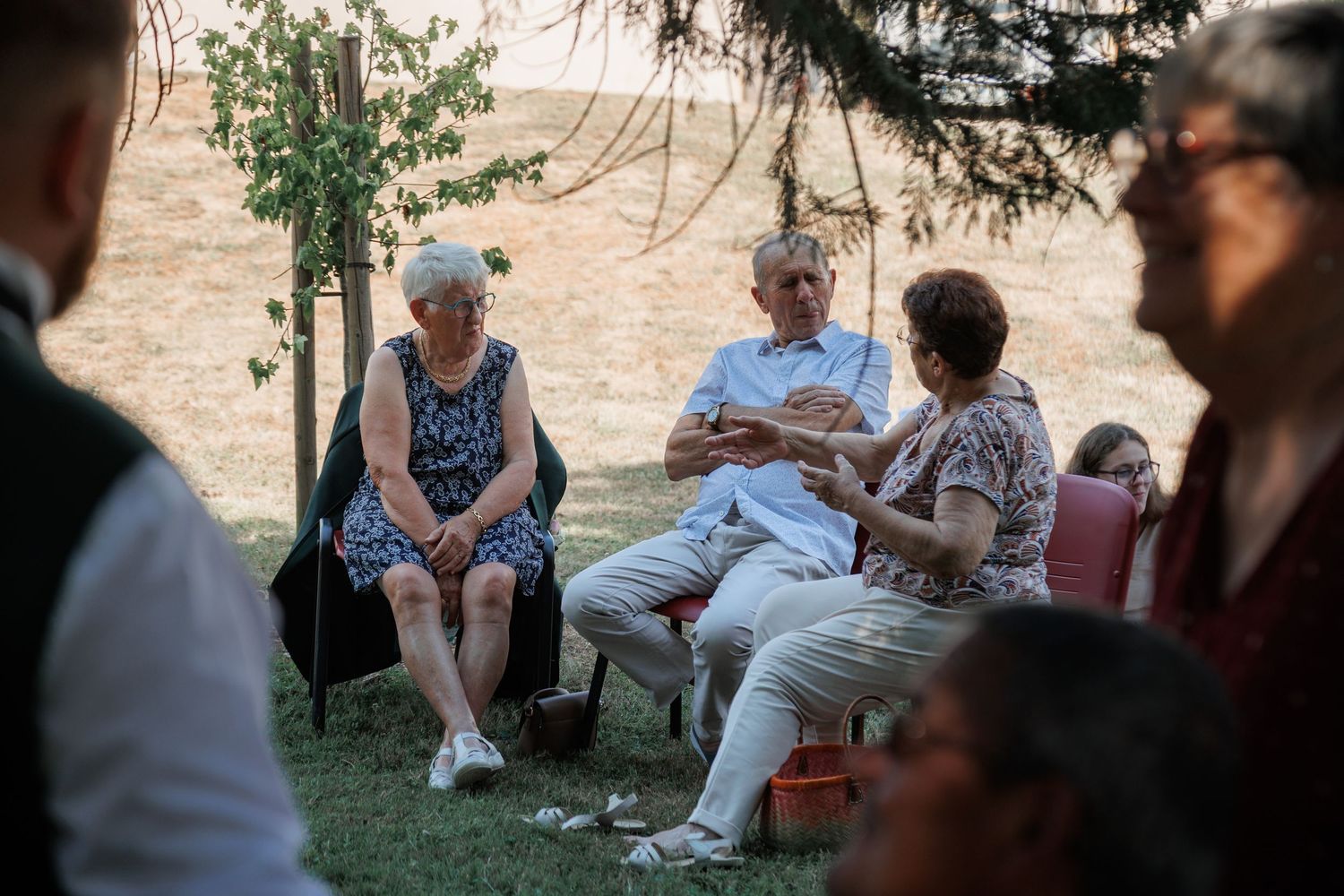 Three elderly people sitting on chairs outdoors at a garden gathering, engaged in conversation.