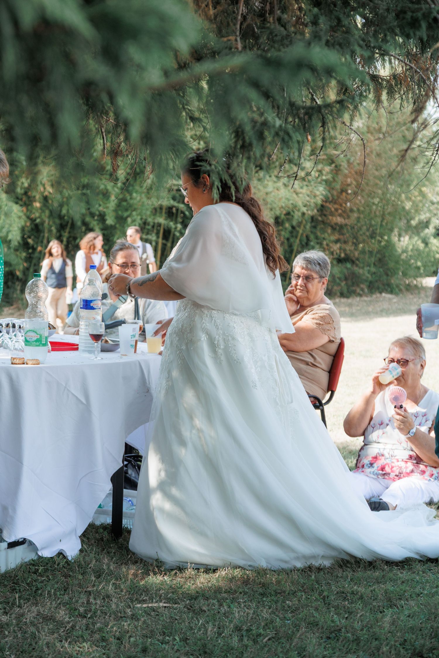 Bride in white lace gown stands at outdoor reception table surrounded by seated guests under trees.
