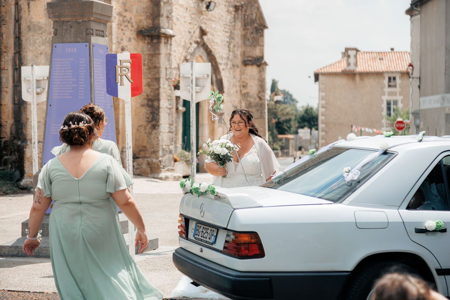 Bride in white gown holds bouquet near decorated car outside French town hall on wedding day.