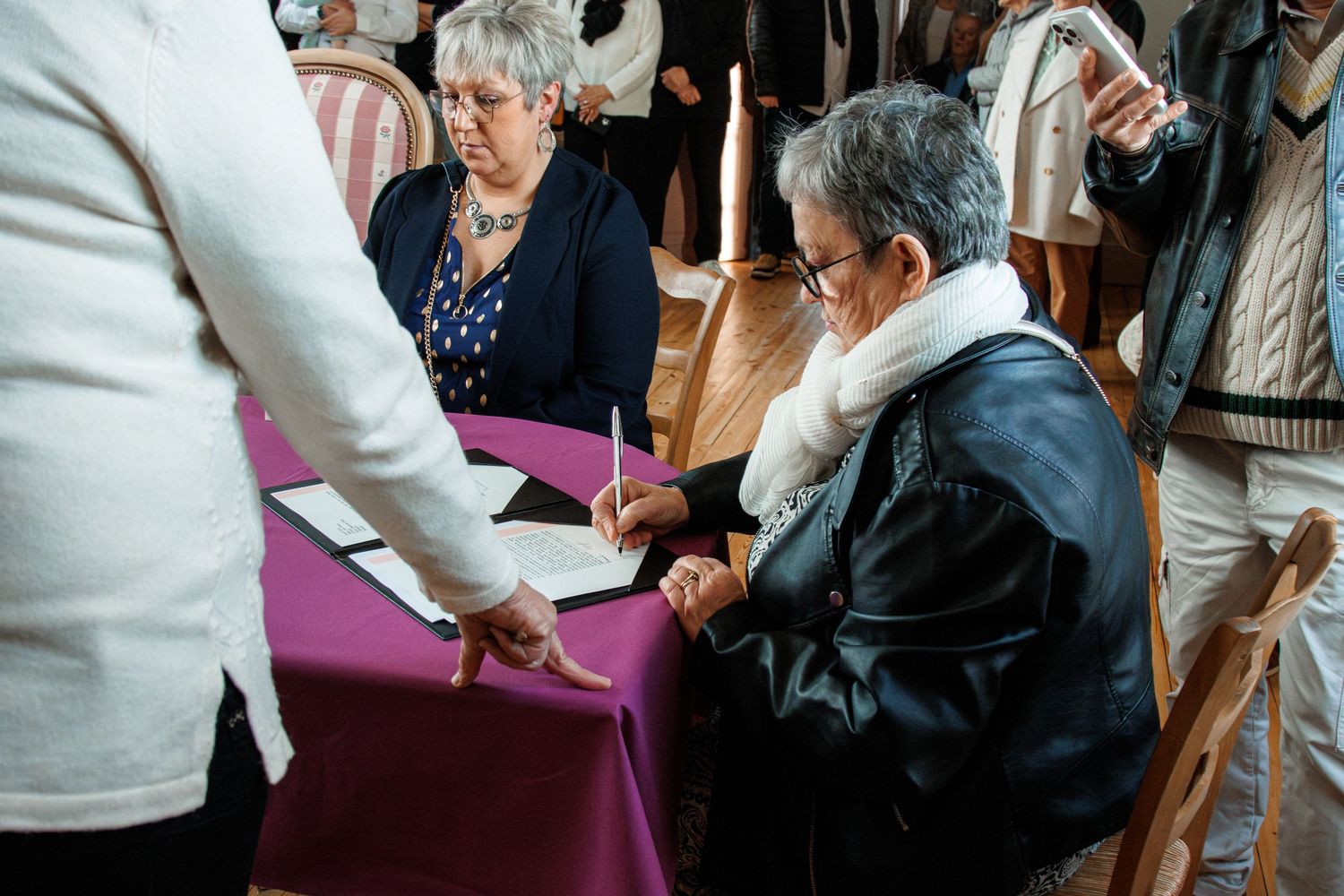 People signing official documents at a formal ceremony table with purple cloth in an elegant room.